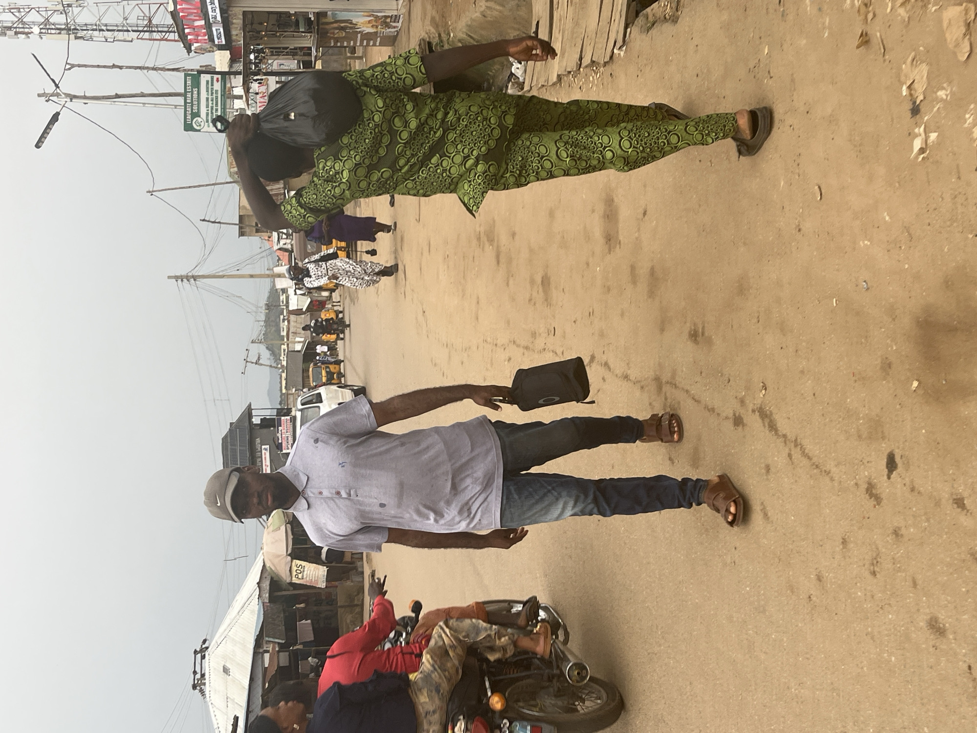 The image depicts a street scene in Abuja, Nigeria, on an overcast day. In the foreground, a man wearing a grey polo shirt, blue jeans, and brown sandals walks towards the camera, carrying a black bag in his right hand. To his right, another man in a green patterned traditional attire with a black bag slung over his shoulder walks away from the camera. Behind them, a person on a motorcycle wearing a red shirt and camouflage pants is visible on the left. The street is unpaved and dusty, with various stalls and buildings lining the sides. Electrical wires and a cell tower are visible in the background, suggesting an urban environment. Several vehicles, including yellow auto-rickshaws, are also present on the street.