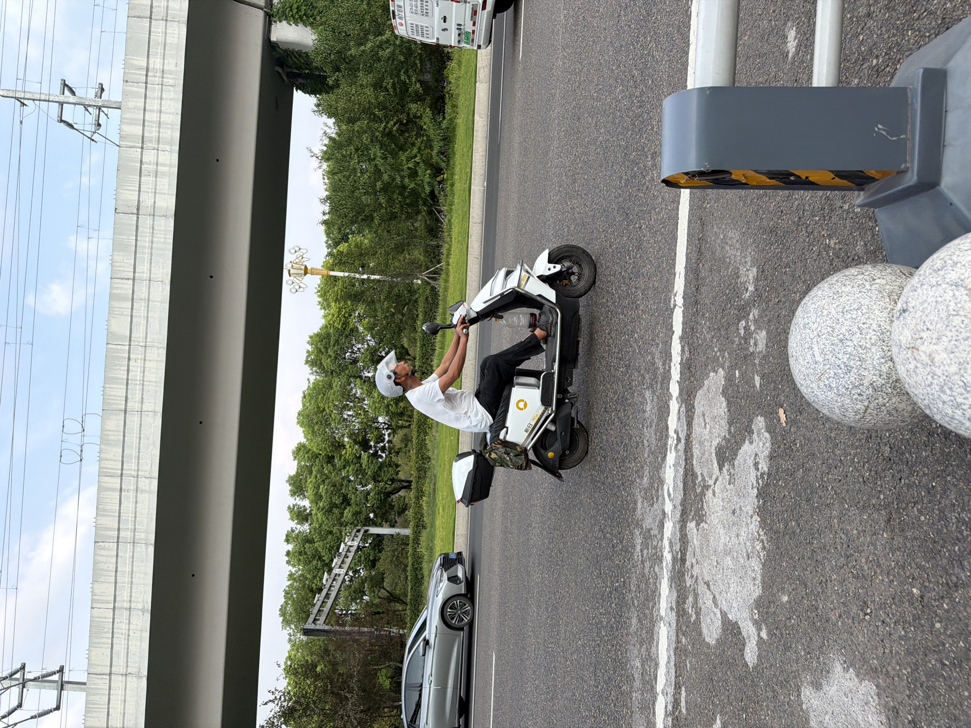 The image shows a man riding a white electric scooter on a road in Jiangjin, China. He is wearing a white t-shirt, dark pants, and a white helmet. The scooter has a black storage box on the back, and a camouflage bag is attached to the box. A clear plastic bottle is attached to the side of the scooter. In the background, there is a silver car, trees, a highway overpass, and power lines. A white van is visible on the right side of the image. The road surface is asphalt with white lane markings. In the foreground, there are two round, grey decorative stones and a traffic barrier with yellow and black stripes.