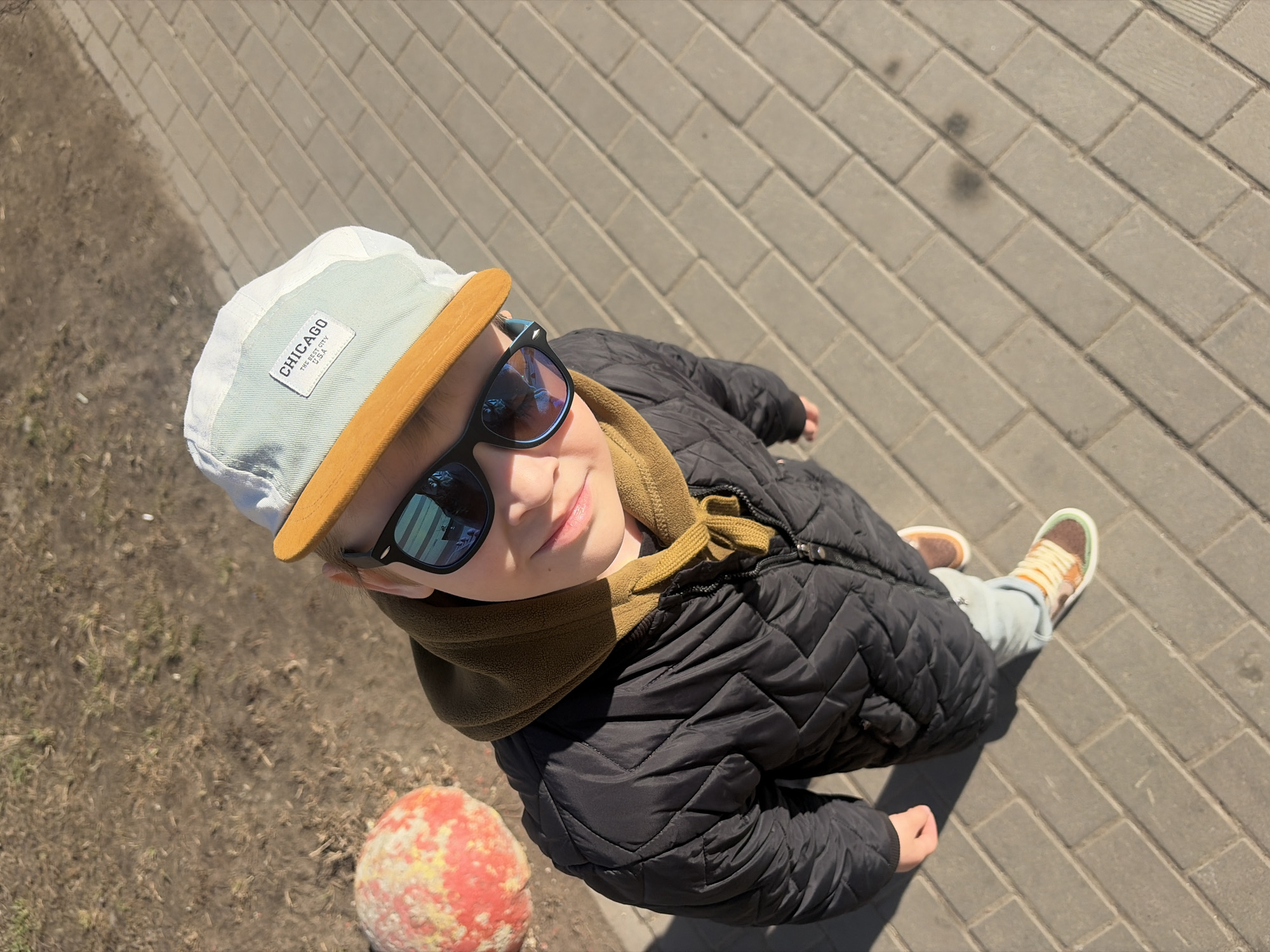 A young boy in a black quilted jacket, brown hoodie, sunglasses, and a baseball cap that says "Chicago The Best City USA" is standing on a brick sidewalk. A patch of dirt with what appears to be a painted sphere is visible near the sidewalk's edge. The location is inferred to be Saint Petersburg, Russia, based on details that may be recognizable to someone familiar with the city.
