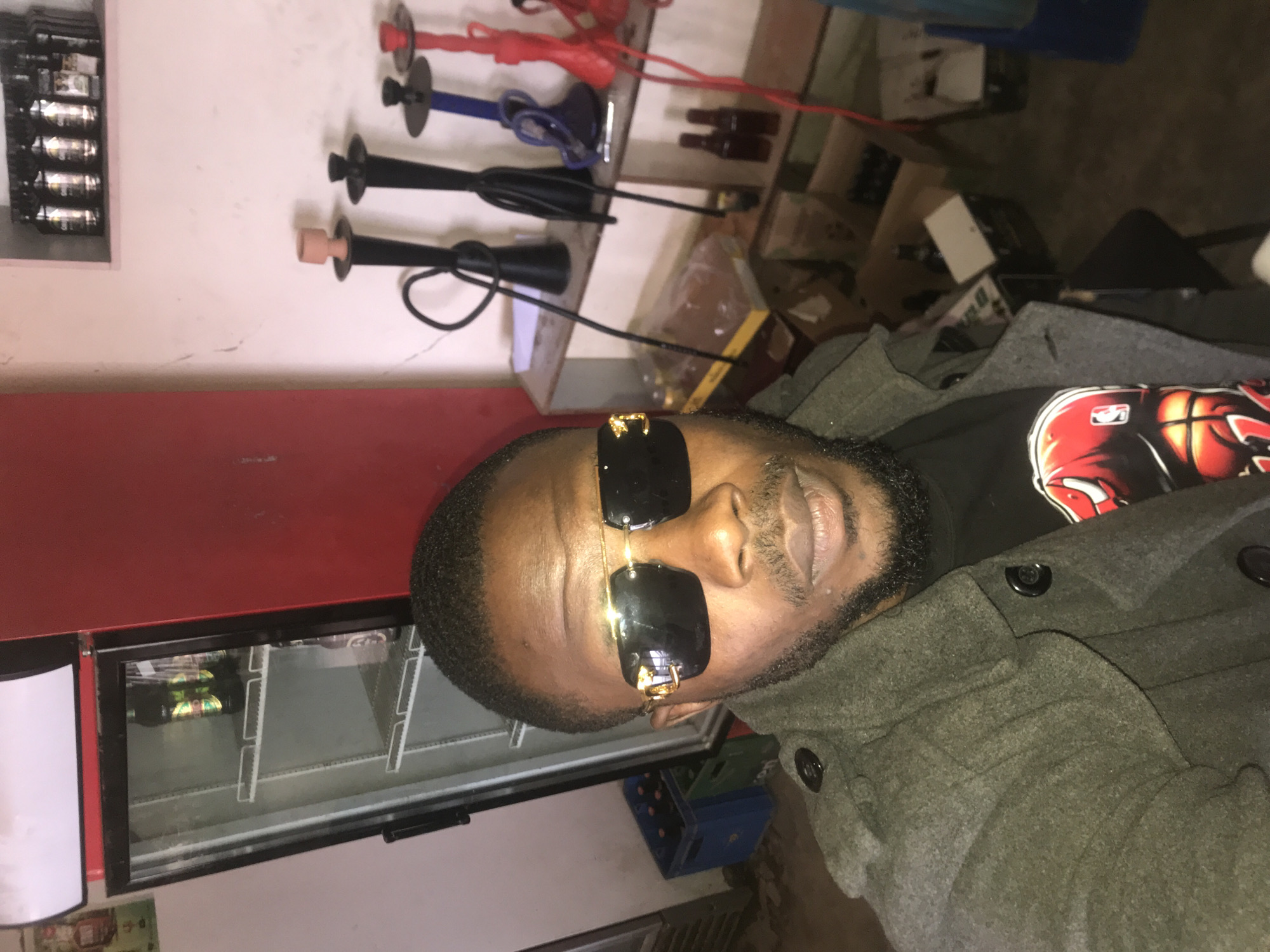 A portrait-style shot features a man indoors, positioned with his head tilted to the right and the image rotated 90 degrees clockwise from its intended upright orientation. The man has dark skin, a neatly trimmed beard, and a mustache. He is wearing large, dark, rectangular sunglasses with gold accents, which reflect a bright light source, obscuring his eyes. He is dressed in a dark grey or olive green long coat with visible large black buttons, over a black t-shirt. A red graphic on his t-shirt shows part of a basketball and a stylized character or logo, with a small NBA logo also visible.
The background is a dimly lit, somewhat cluttered indoor environment. To the man's left (when upright), a red commercial refrigerator or cooler with a glass door is partially visible, containing some items. Above the red appliance, a white wall features a shelf holding several objects resembling hookah pipes with various colored hoses (red, blue, black) and dark stems. To the right of these pipes, small bottles are also present on the shelf. The area further to the right and below the shelf is cluttered with cardboard boxes and other miscellaneous items, suggesting a storage or casual backroom setting. A blue plastic crate is visible near the man's right shoulder. The man appears to be posing for the photograph.