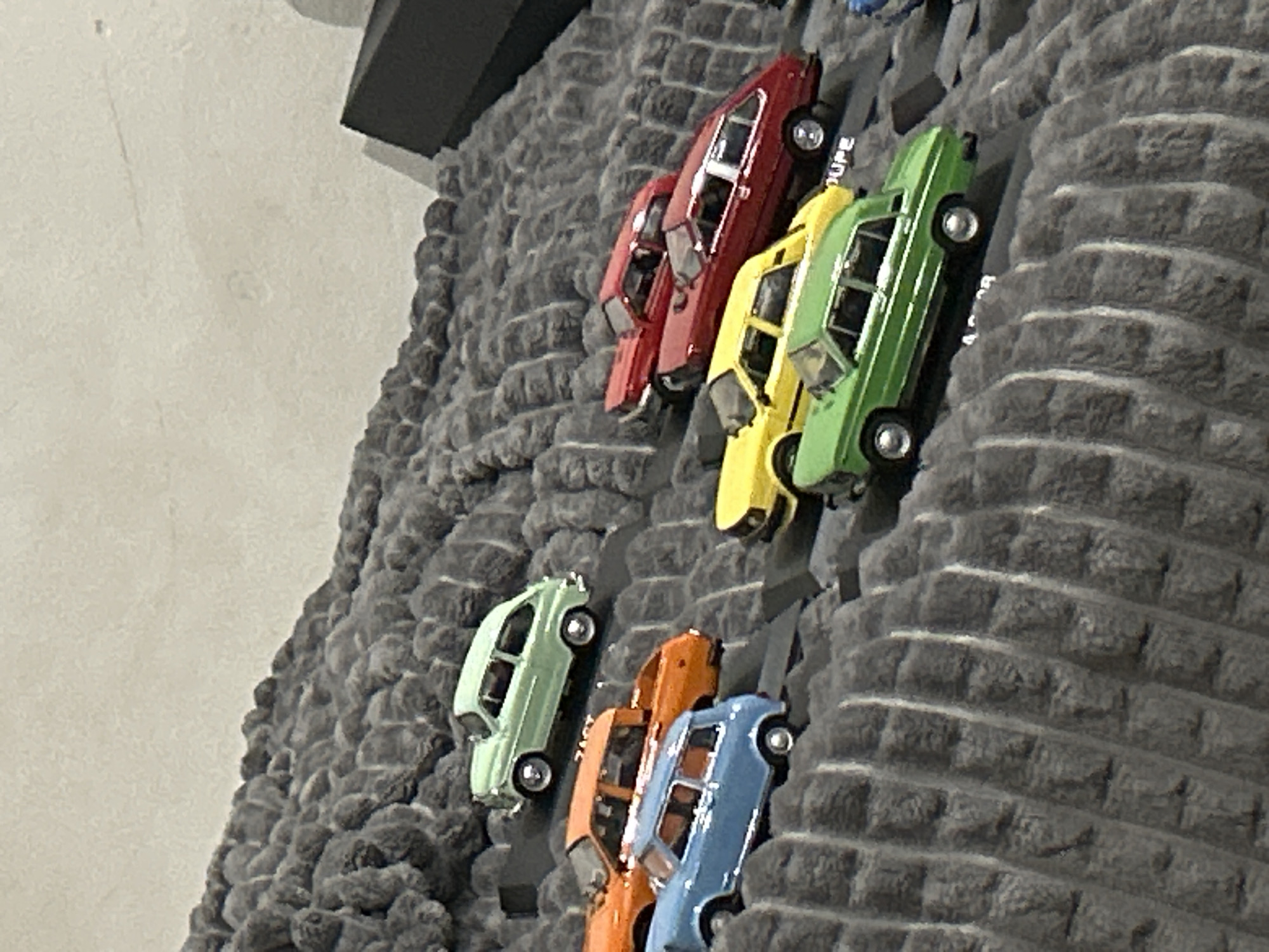 The image shows a decorative wall hanging featuring several miniature cars, each a different color (red, yellow, green, light green, orange, and blue). These toy cars are affixed to a textured, gray, brick-like surface that likely represents a wall or cliff face. The hanging appears to be mounted on a wall in an indoor setting, with a white wall visible to the left. A dark gray or black structural element is visible near the top of the image. The cars are arranged diagonally, suggesting a climbing or descending motion.