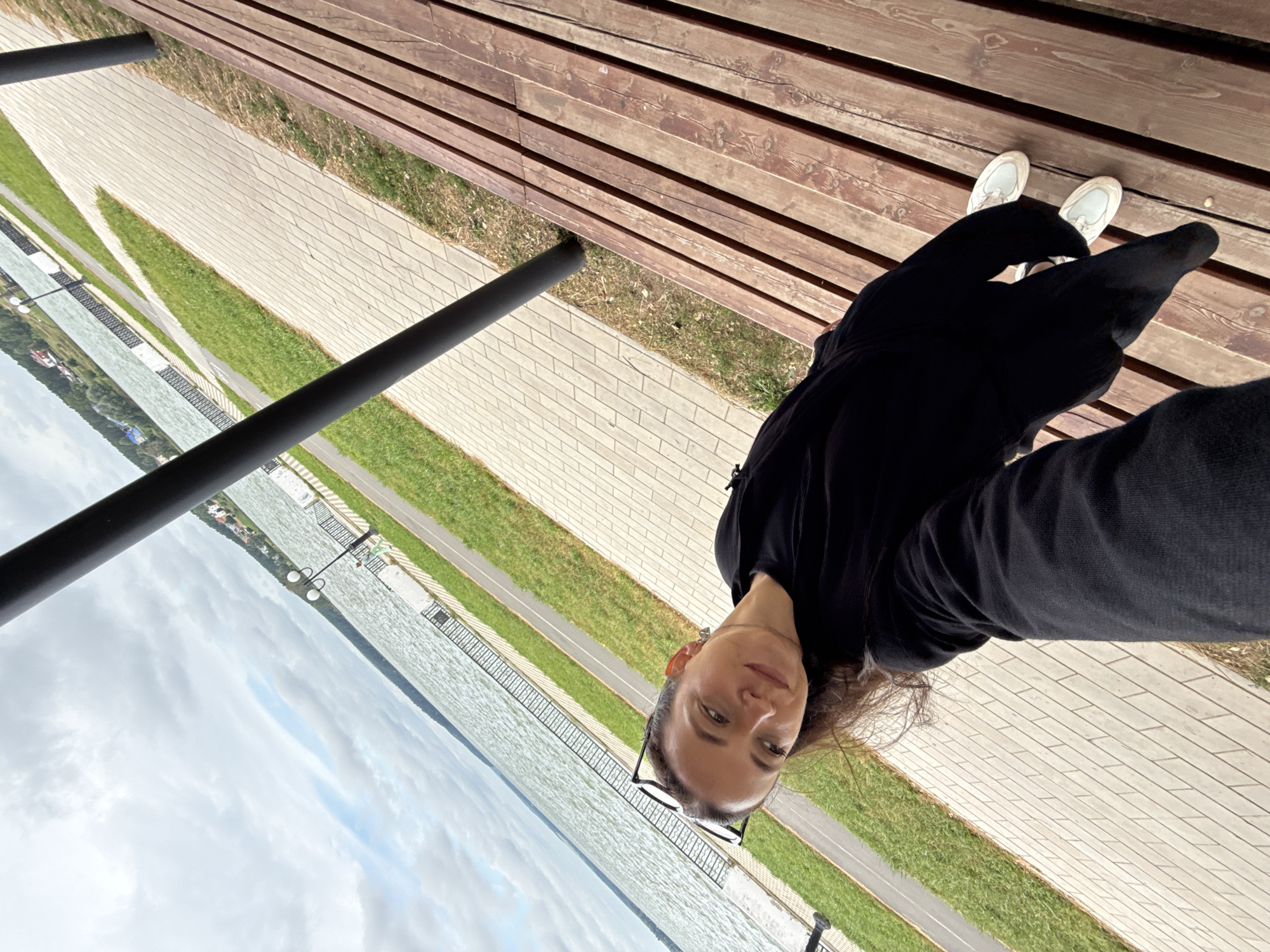 A woman in a black top and pants, wearing white sneakers, is seated on a wooden bench and taking a selfie. She is outdoors in Votkinsk, Russia, with a body of water and a tree-lined shore in the background. A paved walkway and a patch of grass are visible in the foreground. The sky is overcast.