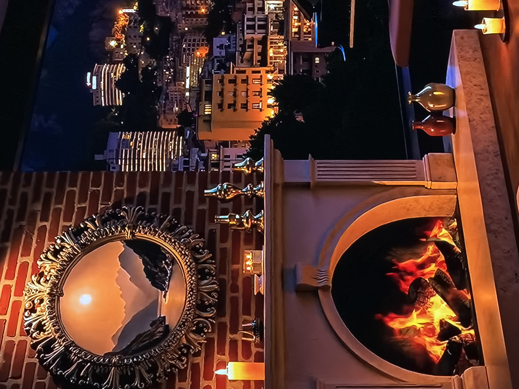 The image, which is rotated 90 degrees clockwise from its intended orientation, showcases an opulent indoor setting with a nighttime city view.
Through a large window (visible at the top of the correctly oriented image), a vibrant city skyline at night is displayed. Numerous buildings are illuminated, their windows glowing with yellow and white light against a dark sky, hinting at the urban landscape of Sofiivska Borshchahivka, Ukraine.
The interior features a prominent fireplace structure. Within its arched opening, artificial logs burn with realistic, bright orange and yellow flames, creating a warm focal point. Above the fireplace, on a mantelpiece, several lit candles cast a soft glow. Two small, decorative vases, one green and one reddish-brown, are also placed on the mantel.
On an adjacent brick wall (to the left in the corrected orientation), an ornate circular mirror with an intricate golden frame hangs. The mirror's reflection uniquely captures a serene scene of a bright, round sun low in the sky, possibly a sunset or sunrise, over what appears to be a mountainous or cliff-filled landscape.
Additional lit candles are visible throughout the foreground, contributing to the cozy and inviting atmosphere. Decorative gold-colored elements are also present on the wall between the mirror and the fireplace. The overall ambiance is one of warmth, luxury, and comfort, blending interior richness with views of both a bustling city night and a tranquil natural scene. There is no visible text in the image.