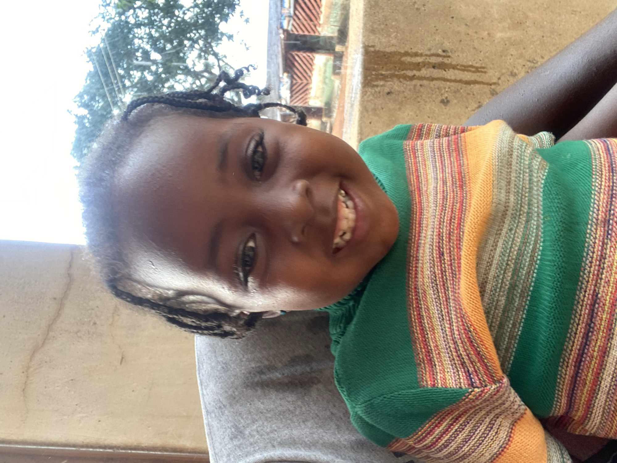 A young child with dark skin and braided hair is smiling broadly at the camera. They are wearing a green, yellow, and red striped sweater. The child appears to be sitting on someone's lap, who is wearing gray clothing. The background includes foliage and a building facade. While the location is said to be Kuru Dungus, Nigeria, there is nothing to explicitly confirm this in the image.