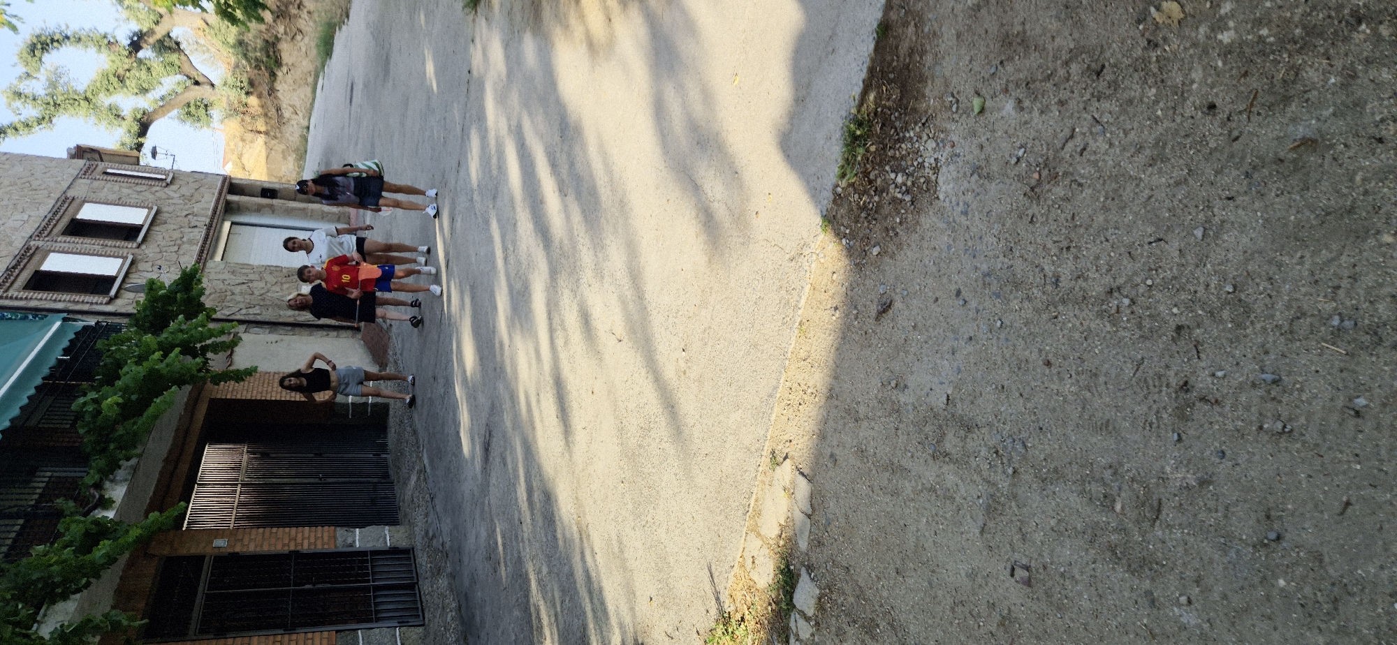 Six individuals are walking on a paved street in front of a two-story building. The building has a stone facade and a dark green awning. The individuals are a mix of adults and children, and they are all casually dressed. One child in the center is wearing a red shirt with the number 10 on it and blue shorts. Another child, a girl, is wearing a black crop top and denim shorts. An adult woman on the far right is wearing a dark skirt and a grey top with a bag over her shoulder. The street is partially shaded by trees and buildings, with shadows cast across the pavement. The surrounding environment appears to be a residential area with some vegetation.