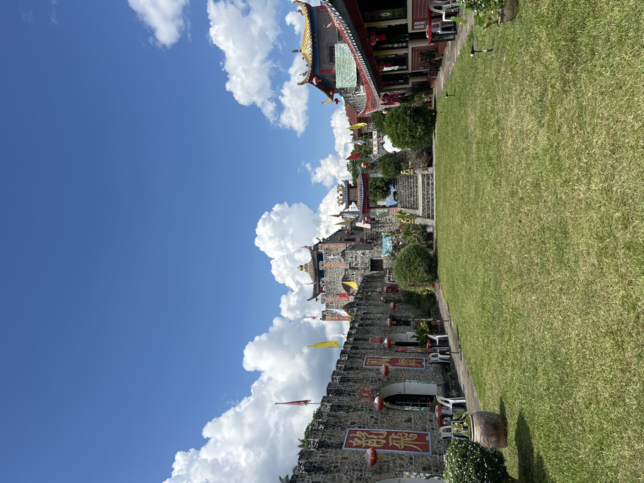 The image displays an outdoor scene at an establishment in Ban Mai Saha Sampan, Thailand, featuring architecture with prominent Chinese influences. A meticulously maintained rectangular lawn of short green grass occupies the foreground and center. To the left, a fortified-style wall constructed of grey, irregularly shaped stones with crenellated battlements extends from foreground to background. This wall features several arched openings, outside of which two white plastic tables and chairs are positioned. Five red traditional Chinese lanterns hang from the wall, and three red banners with yellow Chinese characters are affixed vertically. A large, light brown ceramic pot rests in the lower-left corner.
In the mid-ground and background, a multi-tiered fortress-like structure, also built from the same grey stones and topped with roofs featuring ornate, upturned yellow eaves and a golden finial, stands prominently. Several red and yellow flags are visible on poles around this central building. A stone staircase with decorative railings leads up towards this main structure, passing under an ornate archway with a Chinese-style roof. To the right of the lawn, a building with a red tiled roof and yellow upturned eaves is visible, from which additional red lanterns hang. White plastic tables and chairs are also present on a paved area adjacent to this building. Small, round green bushes are interspersed throughout the landscape. The scene is illuminated by bright sunlight under a clear blue sky dotted with numerous fluffy white cumulus clouds. No individuals are present in the image.