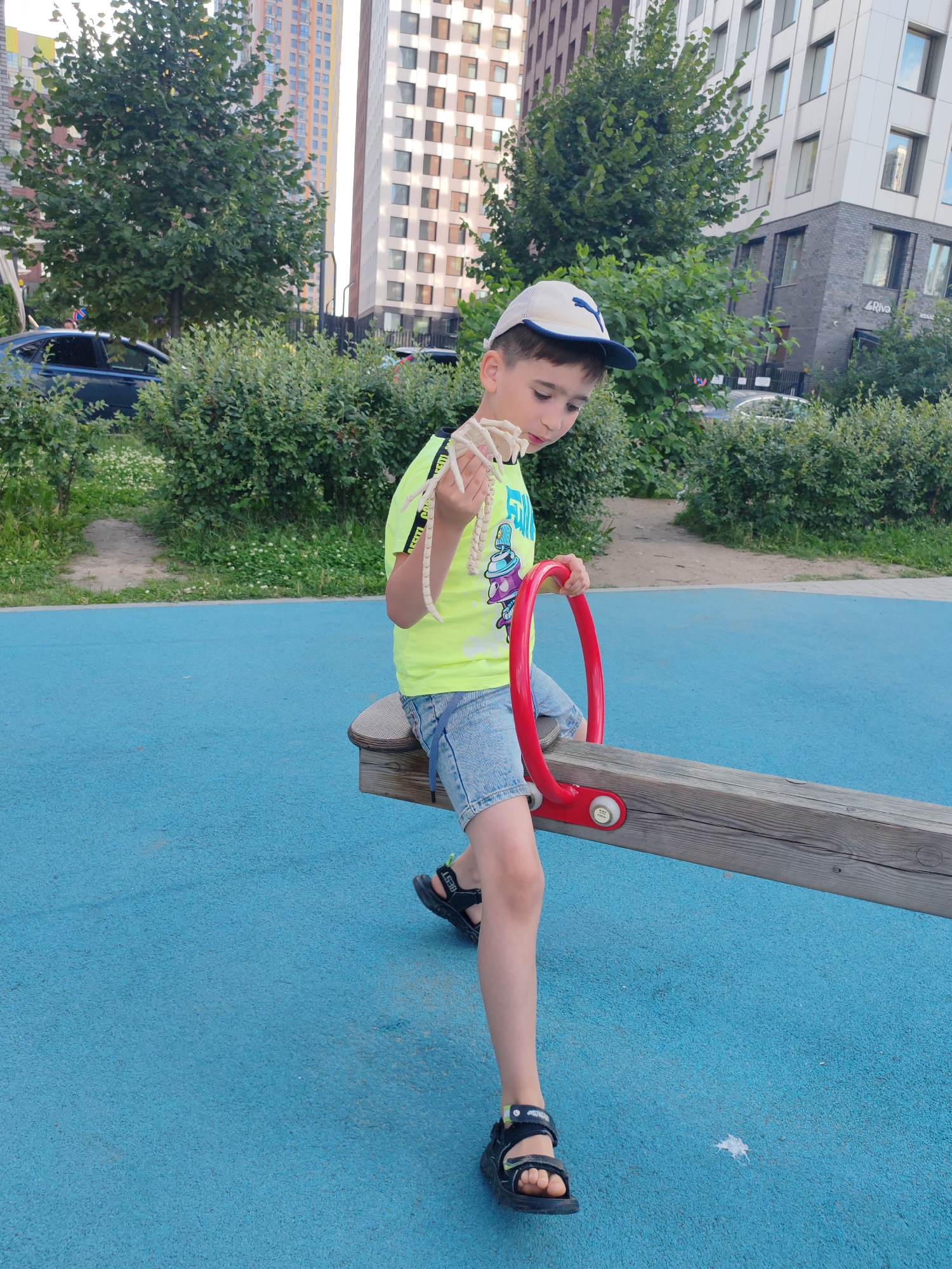The image shows a young boy sitting on a wooden seesaw with red handles on a bright blue playground surface. He is wearing a lime green t-shirt with black and yellow accents on the sleeves, denim shorts, a white baseball cap, and black sandals. The boy is holding a light-colored toy, possibly a dinosaur skeleton, and looking down at it.
In the background, there are green trees and bushes, and tall modern apartment buildings. One building has a facade with a grid of windows and a white vertical strip. Another building appears to be made of darker brick. The setting suggests an urban park or playground.
The time of day appears to be daytime, possibly late afternoon, given the lighting. The weather seems to be pleasant and sunny, with shadows visible on the ground. The boy's expression is one of focused interest in his toy. There is some visible text on the boy's shirt which is part of a graphic and on his sandals. The word "BEST" is visible on the strap of his right sandal. The location context is Moscow, Russia.