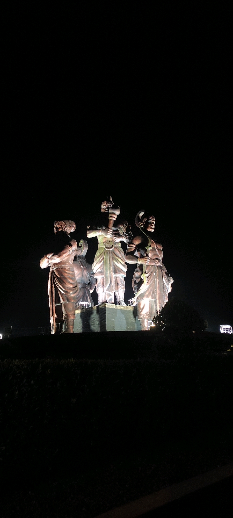 In Purwodadi, Indonesia, a nighttime scene features a monumental statue of three figures standing in front of a dark background. The statues appear to be made of bronze or a similar metallic material and are illuminated from below, casting dramatic shadows. The central figure, the tallest of the three, holds a large club or mace. To its left, a figure with a distinct headdress stands with its arms crossed. To the right, another figure sports a curved headdress and holds a long, thin object, possibly a spear or staff. The statues are positioned on a raised platform, and the foreground is dominated by dark, low-lying vegetation, obscuring the lower portions of the statues. In the distance, on the right side of the frame, a small, brightly lit structure, possibly a building or monument, is visible against the dark sky.