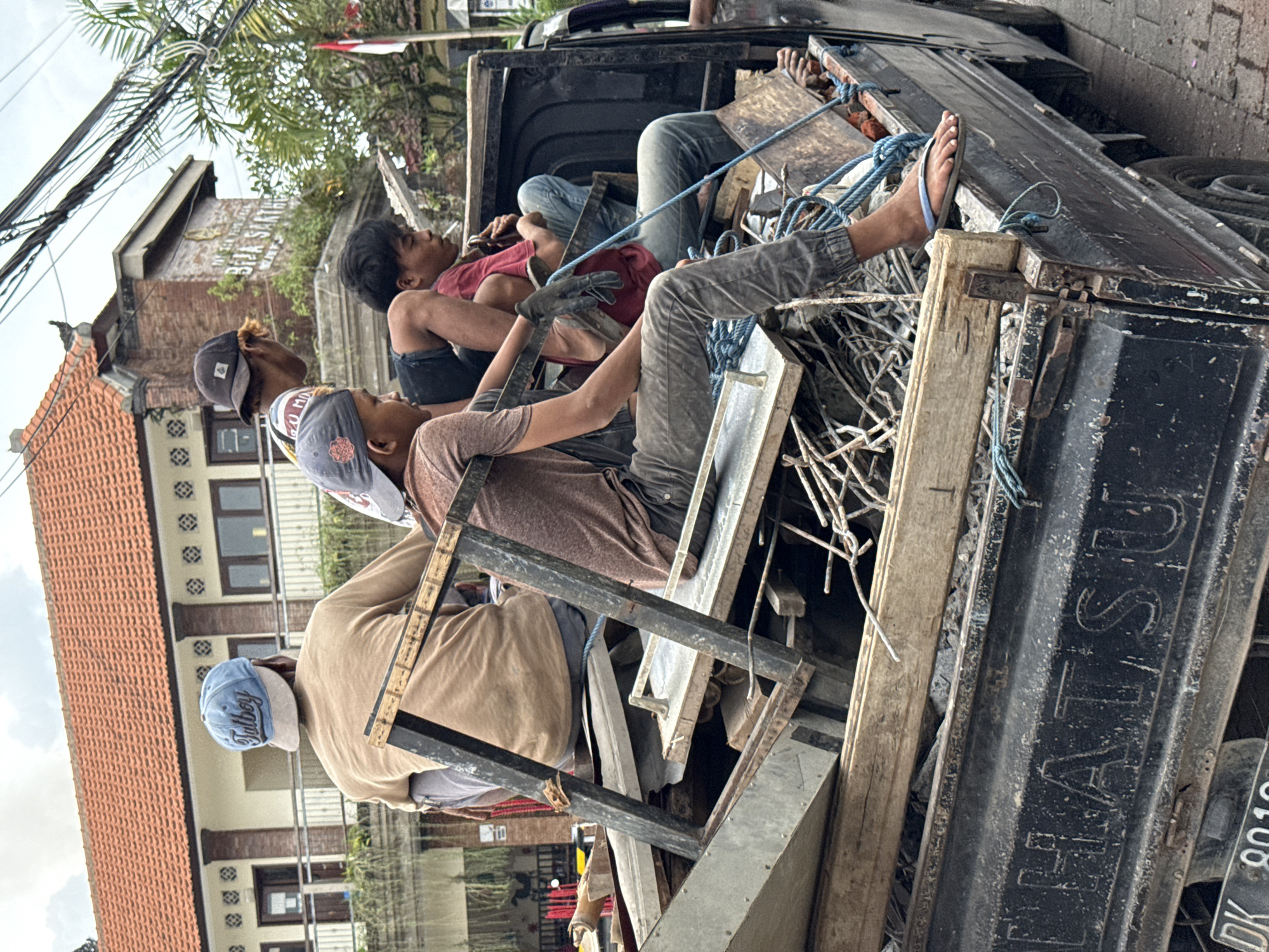 Several men are seen riding in the open back of a black Daihatsu pickup truck, which is heavily loaded with an assortment of scrap materials including metal frames, wires, and wooden planks. The men appear to be resting or in transit, some reclining amongst the debris.
At least four men are visible. One man in a red shirt and dark shorts is lying down. Another, wearing a dark shirt and work gloves, is seated or leaning against the truck's side. Two other men, dressed in light brown and tan shirts, are further forward in the truck bed. One of them wears a light blue baseball cap with "TUTBOY" embroidered on it. Another man has a cigarette in his mouth. Blue ropes are extensively used, seemingly to secure the load and perhaps to provide some stability for the passengers. One man's foot, wearing a light blue flip-flop, is visible resting on a piece of material.
The scene is outdoors, likely during the daytime under an overcast or bright sky. In the background, buildings with distinct reddish-orange terracotta roofs and traditional architecture are visible, alongside lush green foliage and palm trees. Overhead, numerous power lines crisscross the sky. The truck's tailgate clearly displays "DAIHATSU" and part of a license plate, "DK 8019," which is characteristic of vehicles in Bali, Indonesia. The overall impression is one of workers being transported with their collected materials, perhaps after a day of work.