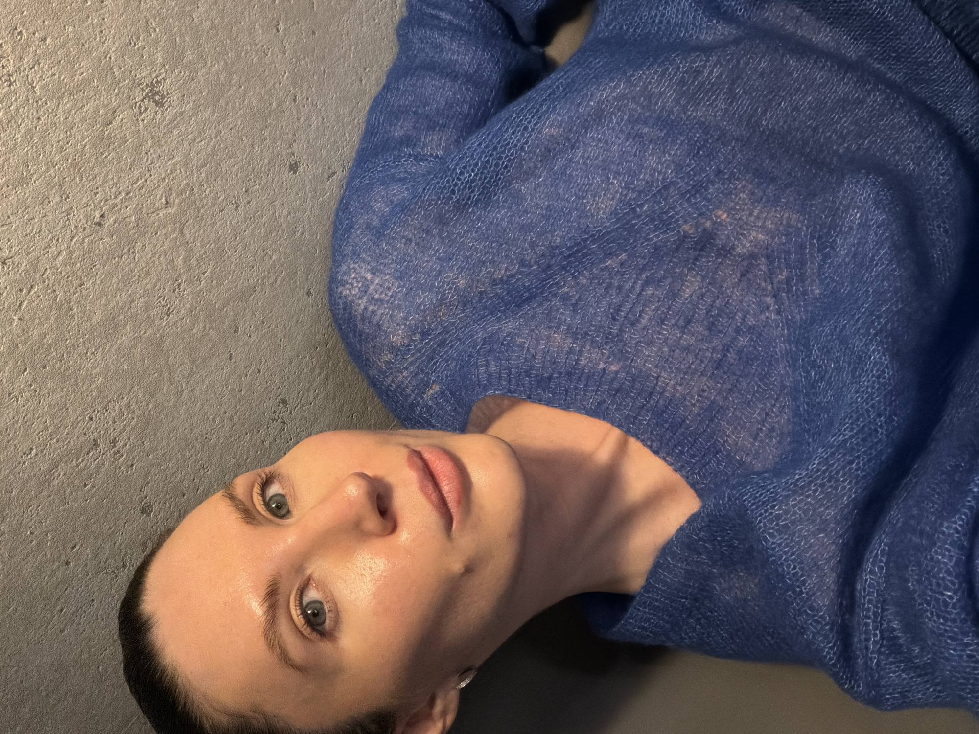 A person is lying on a gray surface, possibly concrete or a similar material. They are wearing a blue, slightly sheer sweater. The individual's face is visible, displaying light skin, light-colored eyes, and dark hair pulled back. The image has no obvious indicators of the location being Perm, Russia.
