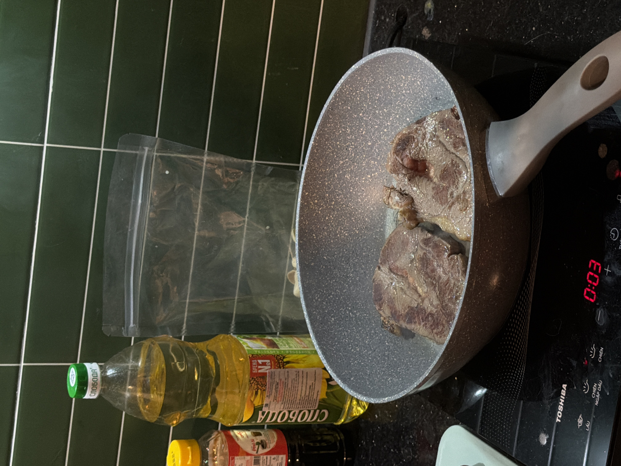 The image captures a close-up view of two pieces of raw steak being seared in a gray, speckled frying pan. The pan rests on a black induction cooktop, which displays a timer reading "0:03". To the left of the pan, a large bottle of sunflower oil with a green cap is visible, along with a smaller bottle with a yellow cap containing a dark liquid. Behind the oil bottle, a clear plastic zipper bag is present. The background consists of dark green tiled walls. The scene appears to be set in a kitchen environment, possibly in Đà Nẵng, Vietnam, given the text on the cooktop display. There are no people visible in the image, and the focus is on the cooking process.