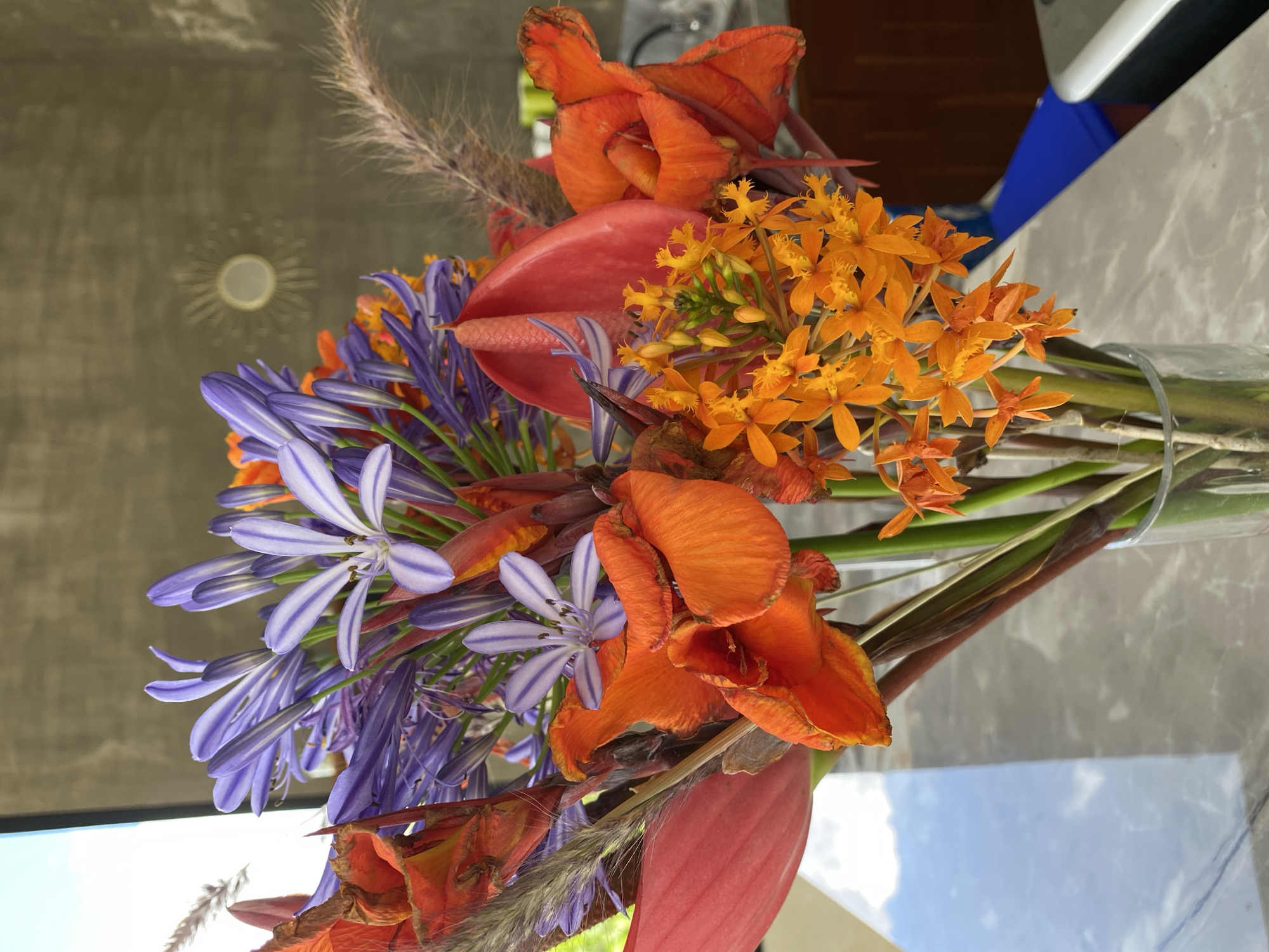 A close-up shot captures a vibrant bouquet of flowers in a glass vase, placed on a reflective marble-patterned surface. The arrangement features a striking mix of colors and textures. Dominating the bouquet are clusters of deep purple Agapanthus flowers, their delicate petals striped with white. Interspersed among them are fiery orange blooms, possibly Anthurium or Heliconia, with their bold, waxy spathes. Smaller clusters of bright orange, star-shaped flowers, likely orchids, add a further burst of color. Adding a touch of softness and texture are feathery, light brown ornamental grasses. The background is dimly lit, with a textured gray wall. A subtle circular reflection, possibly a decorative mirror or light fixture, is visible on the wall. To the left, a bright, out-of-focus area suggests natural light and possibly a window. The overall scene is well-lit, with the flowers taking center stage against a subdued backdrop.