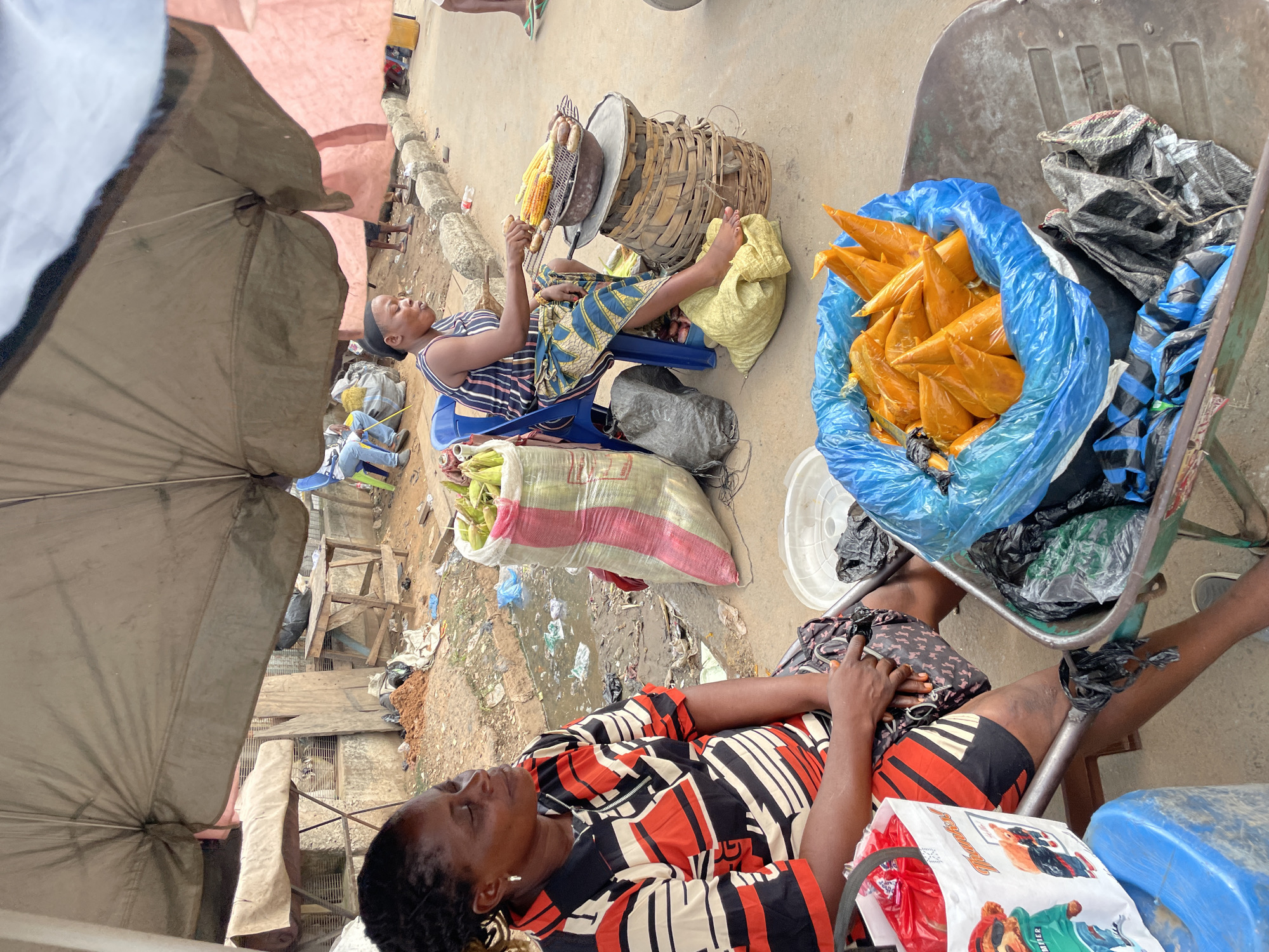 An outdoor market scene shows two women selling food under a large, weathered umbrella. The woman on the left sits in a chair wearing a black, red, and cream striped shirt and a patterned skirt. Beside her is a blue plastic stool with a white bag with images of teddy bears. In front of her is a green wheelbarrow filled with items wrapped in blue and black plastic bags, and a blue plastic container. To her right, a large sack of corn is leaning against a blue chair. The woman on the right sits in the blue chair, wearing a striped top and a blue and yellow patterned skirt. She is grilling corn and sausages on a metal rack over a fire. The grilled items are placed on a woven basket. A large sack of corn is to her left. The ground is dirt and asphalt, with some debris scattered around. In the background, there are tables, chairs, and more vendors. The overall scene depicts a bustling marketplace environment.