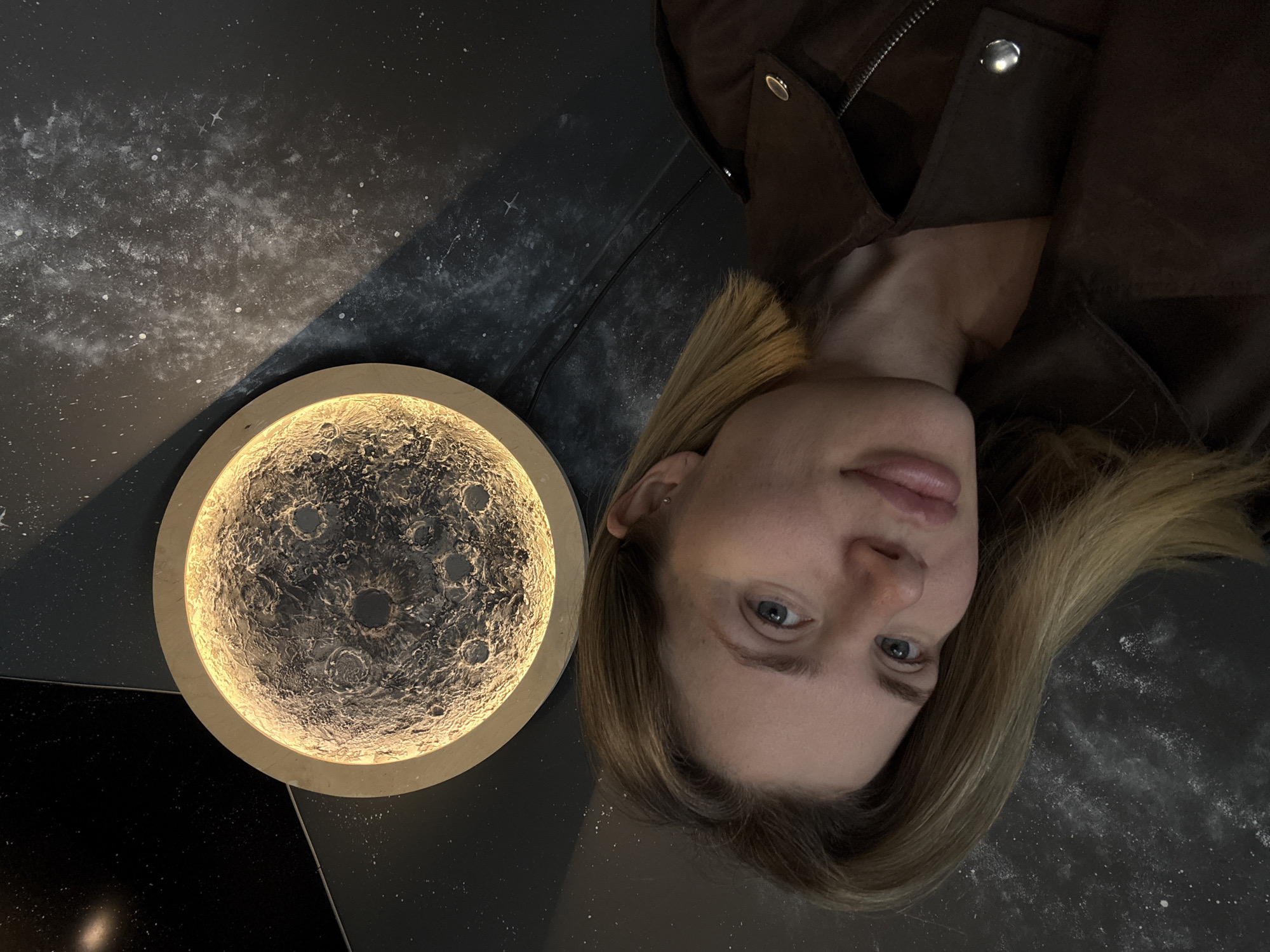 The image captures a woman with fair skin and blonde hair lying on a dark surface that resembles a depiction of space, with painted stars and nebulae. She is wearing a brown jacket. Next to her is a circular light fixture resembling the moon, illuminated with a warm glow, showcasing a detailed texture of craters and lunar surface. The location is presumed to be in Mayma, Russia. The woman is looking directly at the camera.