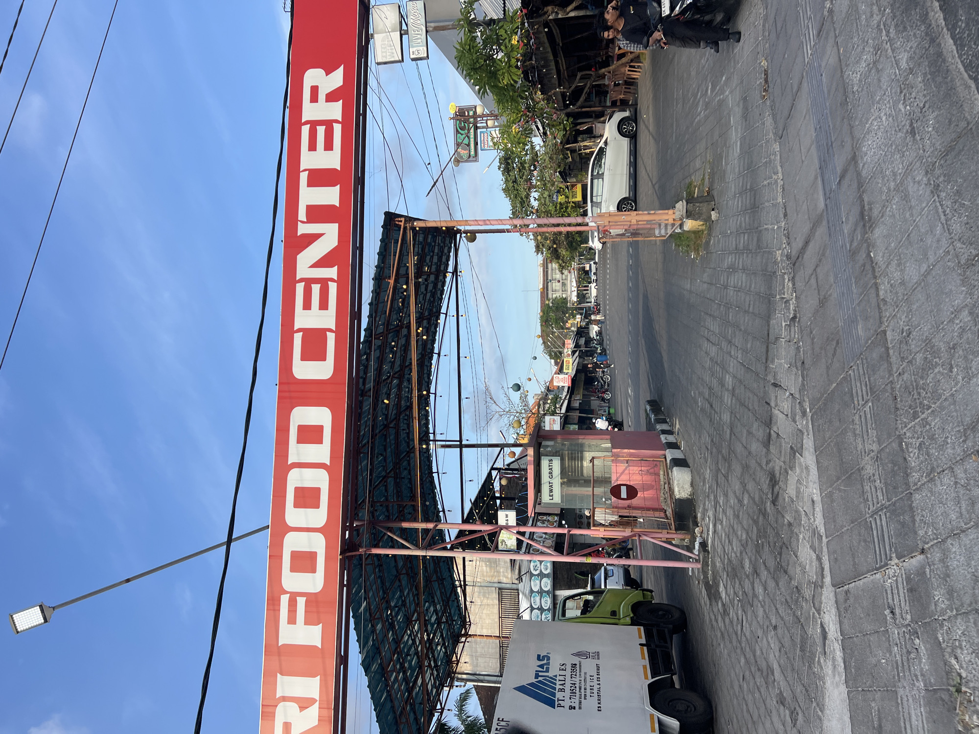 A wide-angle, low-light photograph captures the entrance to the "Kuta Food Center" in Kuta, Indonesia. The iconic red sign with white lettering dominates the upper portion of the image, under a bright blue sky. Below the sign, a covered walkway with exposed metal beams and string lights stretches across the scene, leading into a bustling market area.
On the left, a white delivery truck with "PT. BALI ES" branding is parked. To its right, a pink booth with a "LEWAT GRATIS" (Free Pass) sign and a circular red and white prohibition symbol is manned by an individual. The ground is paved with textured grey tiles, and a black and white striped curb runs along the edge of the road.
In the background, a street lined with food stalls, shops, and restaurants is visible. Several cars and motorbikes are present, with people walking and riding. The overall atmosphere suggests a vibrant and active commercial district. The presence of the "Kuta Food Center" sign and the general architecture and signage indicate that this scene is indeed located in the popular tourist destination of Kuta, Bali, Indonesia.