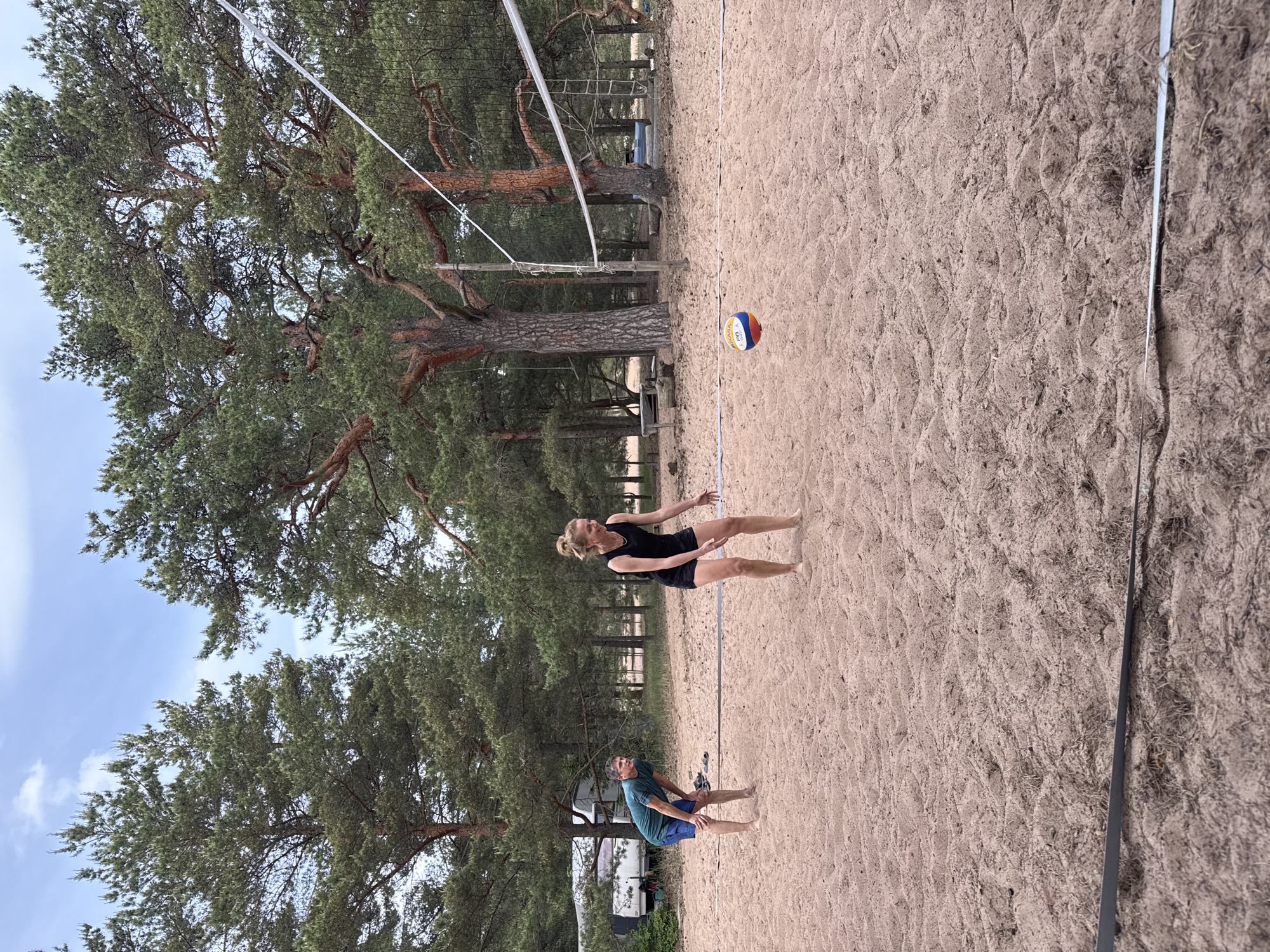 This image captures a beach volleyball court in Zelenaya Roshcha, Russia, set within a sandy area bordered by a pine forest. Two barefoot individuals are present on the sand court. On the right side, a female with blonde hair pulled back, wearing a black tank top and black shorts, is leaning forward with her arms bent and hands open, appearing ready for action, looking towards a multicolored (blue, white, and orange/red) Mikasa FIVB volleyball resting on the sand in front of her, near the net. On the left side of the court, further back, a male with short dark hair, wearing a dark teal t-shirt and dark blue shorts, is bent at the waist with his hands resting on his knees, observing the scene. A white volleyball net spans the court, supported by two wooden posts. The court is delineated by both white and black lines on the sand. The background is dominated by a dense growth of tall pine trees with reddish-brown trunks and green canopies. A partially cloudy blue sky is visible above the tree line. A white camper van or trailer is partially visible among the trees on the far-left side of the background. Additional indistinct structures and objects are scattered within the forested areas behind the court.