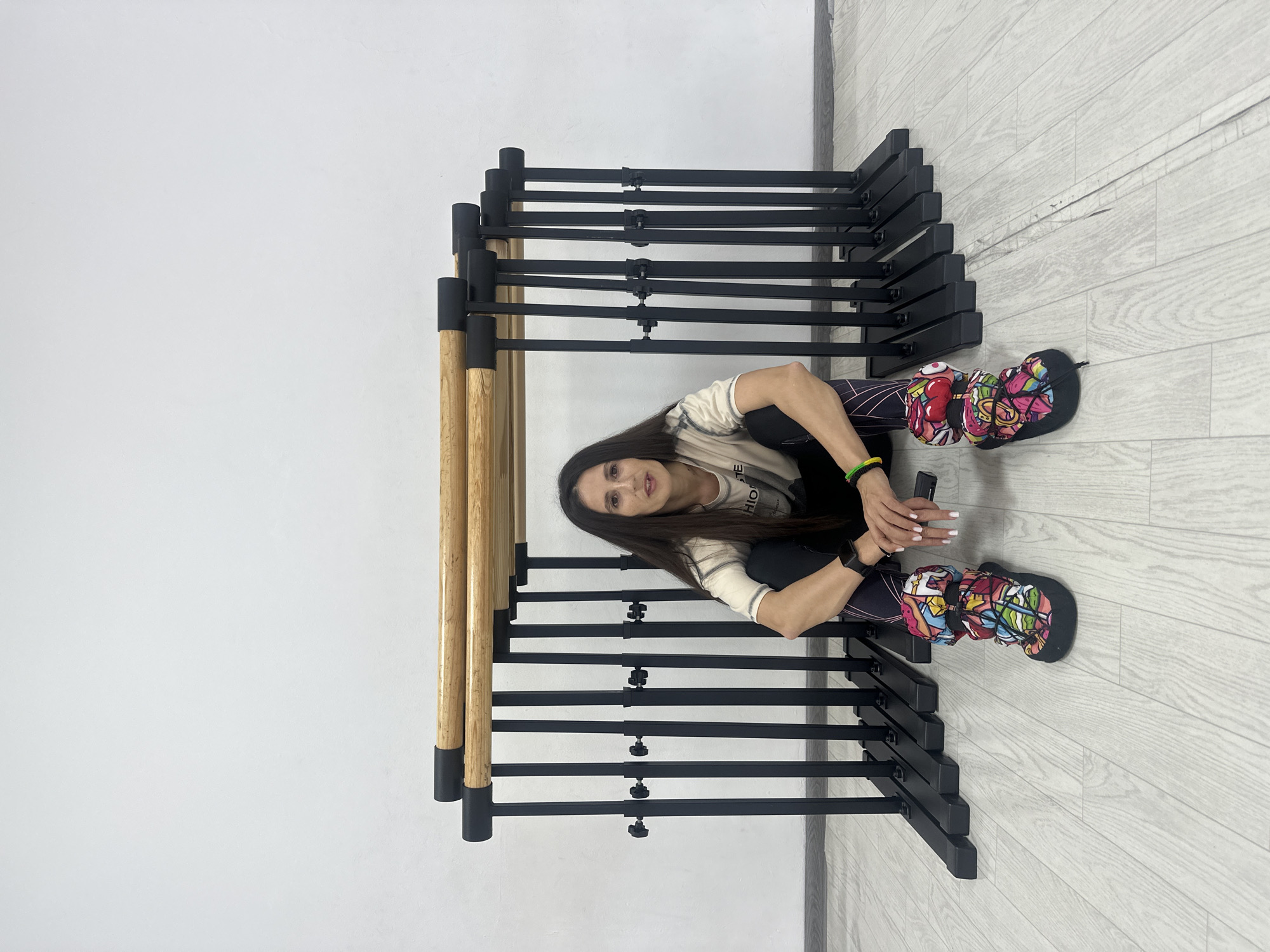 The image depicts a young woman sitting on the floor in an indoor setting in Votkinsk, Russia, surrounded by several portable ballet barres. The room features plain white walls and a light-colored, wood-effect laminate floor.
The woman has long, dark hair and is wearing a light-colored t-shirt with some faint, illegible text, black leggings, and distinctive, multi-colored boots or slippers adorned with a vibrant, graphic design. She is seated with her legs bent, holding a black smartphone in her clasped hands, and looking directly at the viewer with a neutral expression.
The ballet barres consist of black metal frames with light wooden rails. They are arranged in a close, parallel fashion, creating a structured backdrop. The setting suggests a dance studio, fitness area, or a practice space. The lighting is bright and even, indicative of an interior space. No specific time of day or external weather conditions are visible.