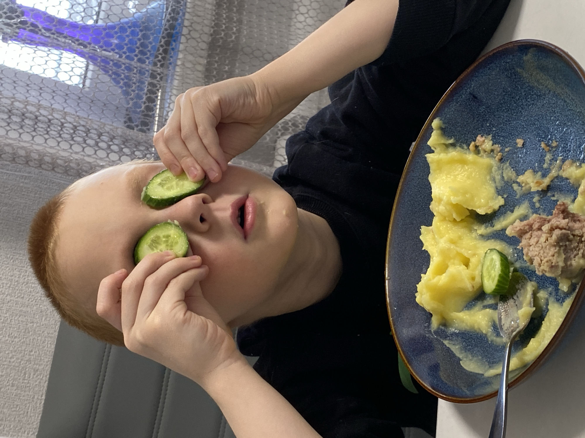 A fair-skinned child with short reddish-blonde hair is holding cucumber slices over their eyes. The child is wearing a black shirt. To the right of the child is a blue oval plate containing mashed potatoes, what appears to be meat, and a cucumber slice. A spoon rests on the plate. The background includes a window with a mesh covering, and a light gray wall. The image was likely taken indoors. There is no definitive indication of location.