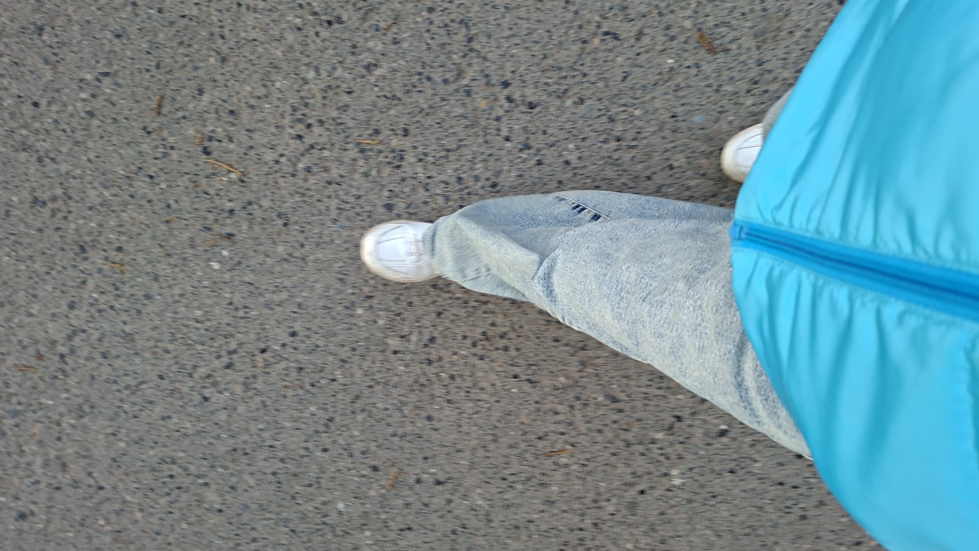 An aerial view shows a person standing on asphalt. The person is wearing light-wash jeans and white sneakers. A bright blue jacket is partially visible on the right side of the frame. The background is gray asphalt. This image appears to be taken from a pedestrian's perspective, potentially while walking. No specific landmarks indicate the location as Chaikovsky, Russia.