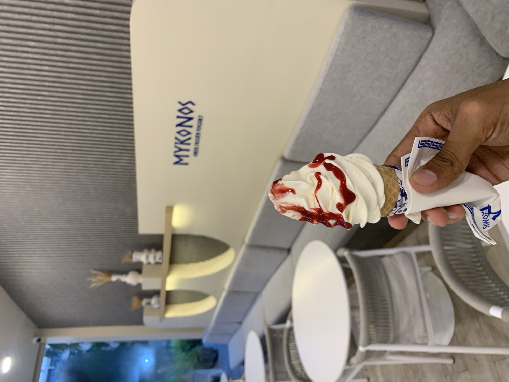 The image, rotated 90 degrees counter-clockwise, presents a close-up of a hand holding a soft-serve ice cream cone inside a modern dessert shop in Pampatar, Venezuela.
The main subject is a white soft-serve ice cream, generously drizzled with a bright red syrup or sauce that drips down its creamy folds. It is served in a waffle cone, partially enclosed by a white paper wrapper adorned with blue patterns and text. A person's hand, with a light brown skin tone, holds the cone.
The background reveals the clean and minimalist interior of the shop. A prominent white wall displays the name "MYKONOS" in stylized blue letters, with "GREEK FROZEN YOGURT" written in smaller text underneath, indicating the establishment's theme and product. To the left, a long, light grey upholstered sofa lines the wall, offering seating. Further back, round white tables and chairs are visible, typical of a cafe setting. Decorative shelving with light-colored vases and dried natural elements adds to the Greek-inspired aesthetic. The ceiling features ribbed grey paneling. Through what appears to be a window or doorway in the distant background, blurred green foliage suggests an outdoor area, possibly indicating late afternoon or early evening lighting.
The ice cream wrapper also features the "MYKONOS" branding and a repeating pattern that resembles Greek characters. The overall impression is that of a contemporary and inviting space to enjoy a sweet treat.