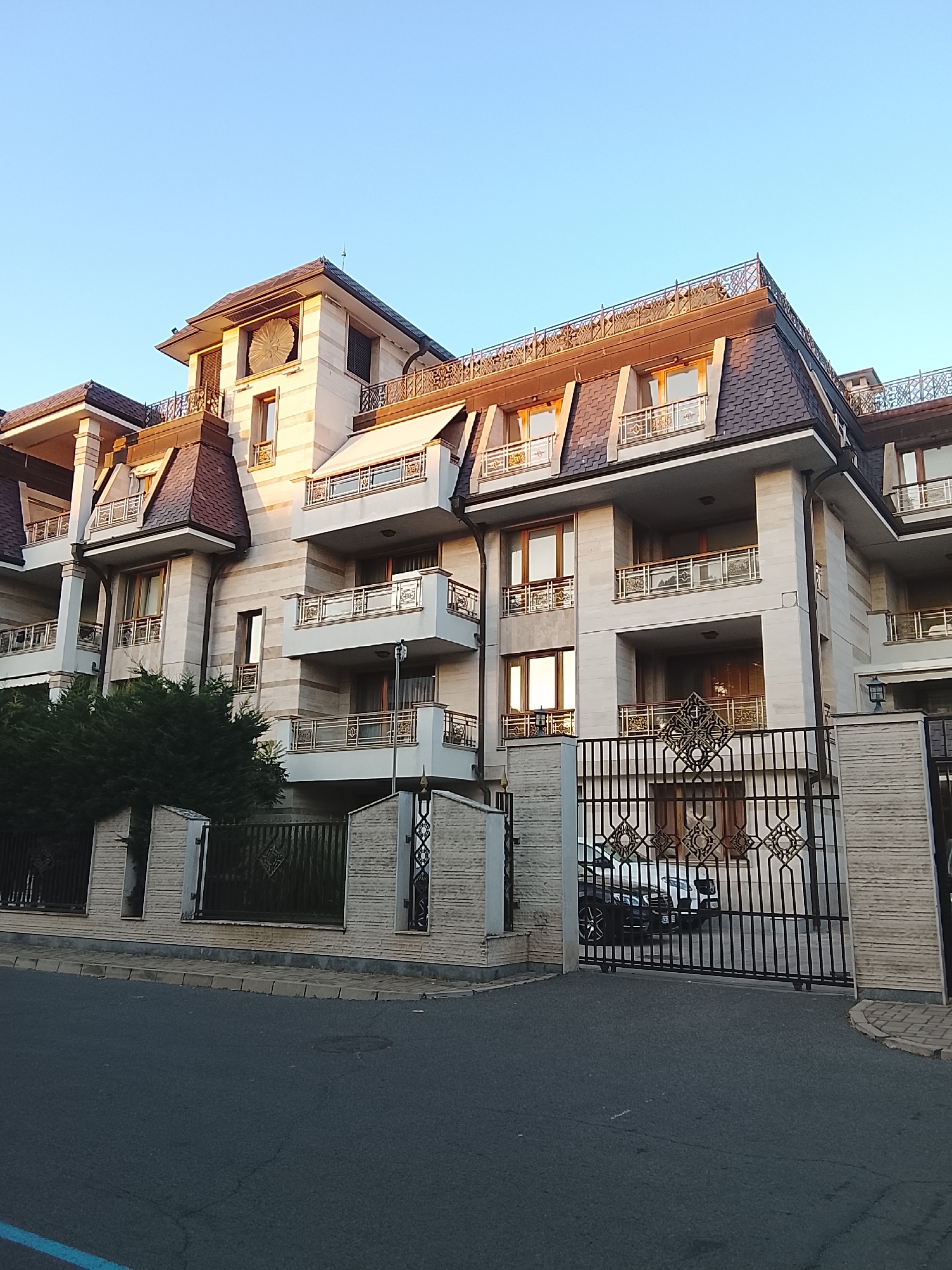 The image depicts a large, multi-story building with a distinctive architectural style. The building is constructed with light-colored stone and features multiple balconies with ornate railings. The roof is dark and has a complex, mansard-like design with dormer windows. A large, decorative metallic gate is visible in the foreground, with a dark-colored car parked behind it. The scene is bathed in the warm light of what appears to be either sunrise or sunset, casting long shadows and highlighting the textures of the building and the gate. The sky is clear and blue. A dense green tree is situated to the left of the building, partially obscuring the lower floors. The road in front of the building is paved, with a sidewalk and a low wall leading up to the gate. The overall impression is one of a grand and well-maintained property. No people are visible in the image. The time of day is clearly indicated by the lighting.