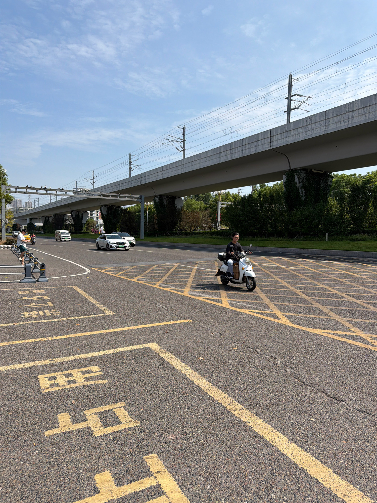 In Jiangjin, China, a person rides a white electric scooter on an asphalt road marked with yellow lines. The scooter has a basket at the front. The rider is wearing a dark long-sleeved shirt and light-colored pants. In the background, a concrete overpass for an elevated railway runs across the scene, supported by pillars. Overhead, power lines stretch across the sky, which is partly cloudy. Beyond the overpass, trees form a dense green line. Other vehicles, including white cars and a white van, are visible on the road. Yellow markings on the road indicate a pedestrian crossing or a no-parking zone. A person in a white shirt and green shorts stands near a metal barrier on the left side of the image, looking towards the road.