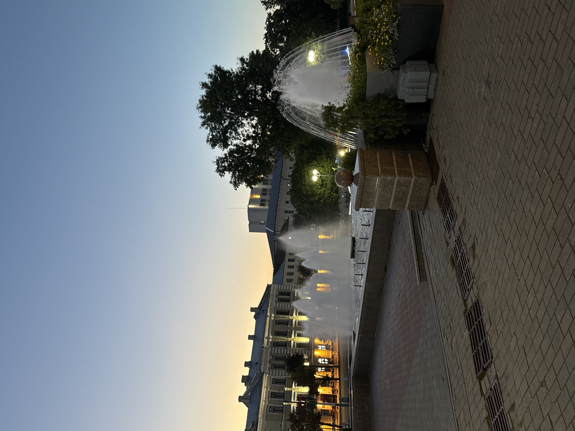 A wide shot captures a public square in Mahilyow, Belarus, at dusk. In the foreground, a brick-paved area features several metal grates set into the ground. A low, stone wall borders a body of water, from which multiple fountains spray water upwards, creating a misty effect. To the right, a planter box overflows with yellow flowers, and a dark green evergreen shrub is planted nearby. A stone pedestal with a spherical finial stands to the left of the planter.
In the midground, a large, mature tree with dark green foliage dominates the right side of the frame. Behind the tree and the fountains, several streetlights illuminate the scene with a warm glow. A modern, light-colored building with multiple windows is visible behind the tree on the left. Across the square, a long, classical building with a light-colored facade and a mansard roof is illuminated by many lights, suggesting it is a public building or office complex. The sky transitions from a pale yellow-orange near the horizon to a clear, deep blue overhead, indicating the end of sunset. The overall scene is peaceful and urban.