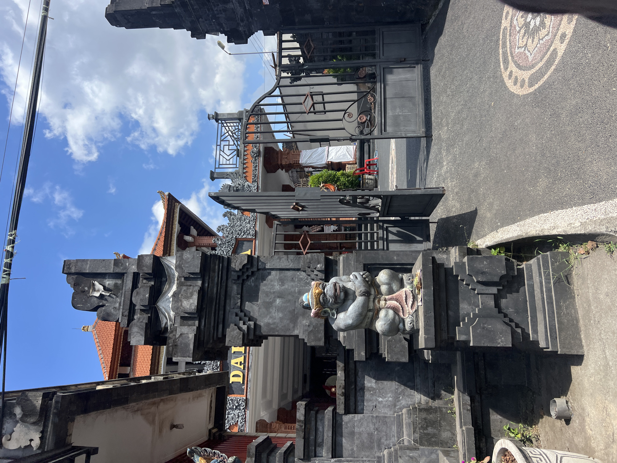 An outdoor shot captures an ornate entrance to a building in Abiansemal, Indonesia. The primary focus is a dark, multi-tiered stone gate adorned with intricate carvings and a weathered stone statue of a creature, possibly a demon or guardian, seated on a pedestal. The statue is gray and wears a colorful headdress. To the right of the statue, a dark metal gate stands open, revealing a paved pathway leading into the property. Beyond the gate, a glimpse of a building with a red-tiled roof and an upper-level balcony with decorative railings is visible. The sky above is blue with scattered white clouds. The ground in front of the gate is asphalt, featuring a circular mosaic inlay. The lighting suggests it is daytime, with shadows cast by the structures.