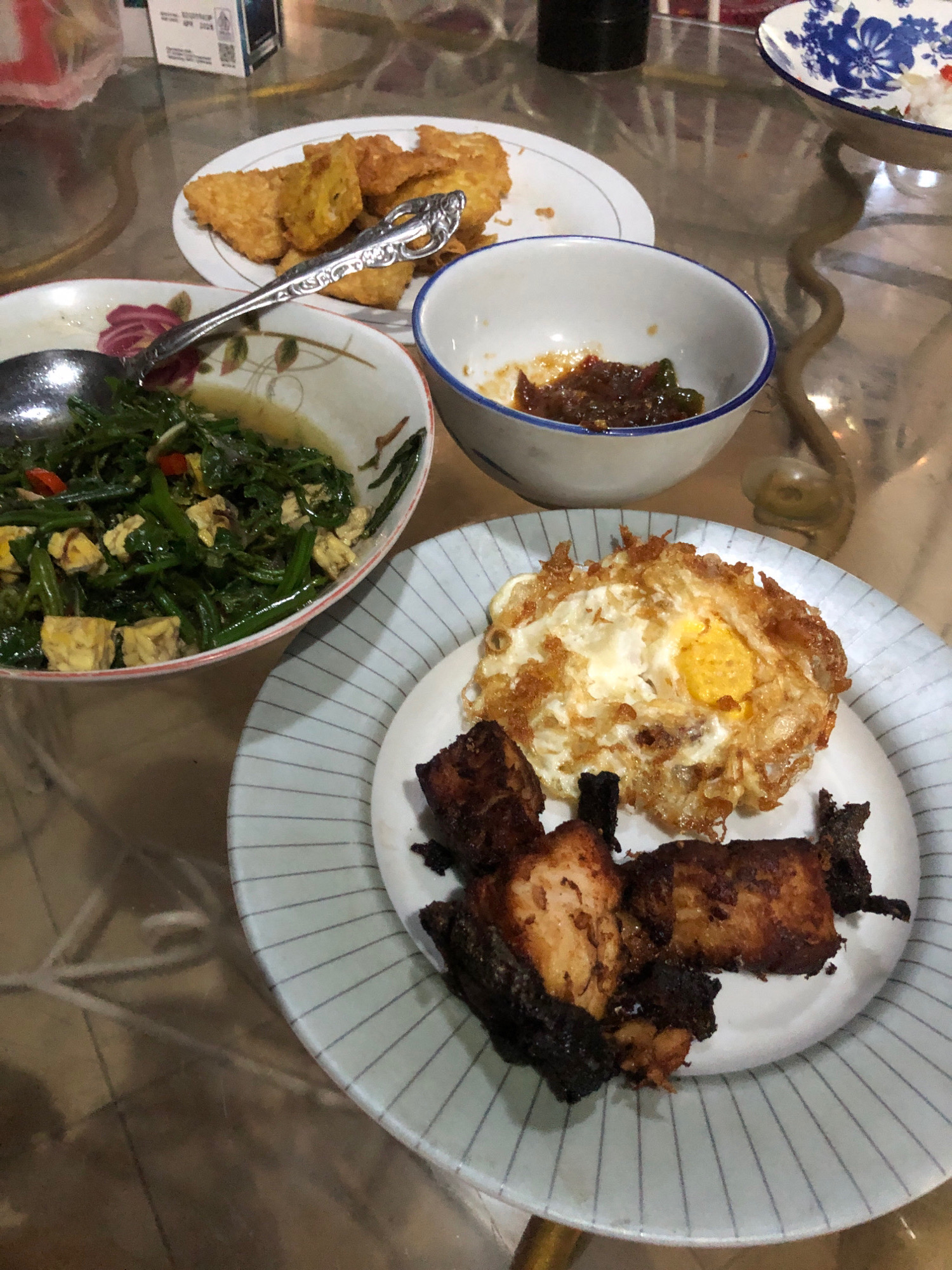 The image displays a table laden with Indonesian food, suggesting a meal is in progress or has recently concluded. In the foreground, a plate holds several pieces of fried fish and a fried egg. To the left, a bowl contains a green vegetable stir-fry with tofu. Behind that, another plate features fried tofu. A small white bowl with chili paste sits to the right of the stir-fry. A blue and white patterned bowl containing rice is visible in the background. The setting appears to be indoors, likely a dining area, with a glass-topped table reflecting the ambient light. The meal consists of traditional Indonesian dishes. The lighting suggests it could be daytime or evening, as the lighting is diffused. No people are visible in the frame. The image evokes a sense of home-cooked meal or casual dining.