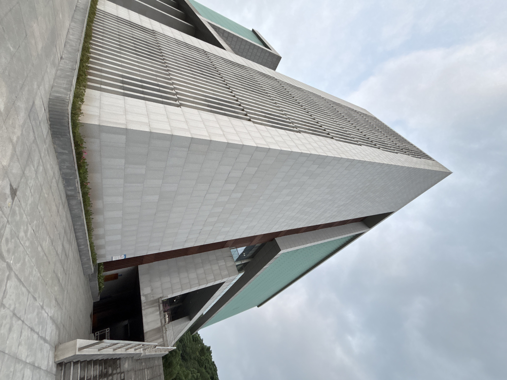 The image shows a low-angle view of a tall, modern building located in Jiangjin, China. The building's facade is primarily white with horizontal lines. The top of the building has angular, cantilevered segments with green roofing. The sky is overcast. A green strip of vegetation sits atop a low retaining wall beside a walkway. The building appears to be the Jiangjin Museum.