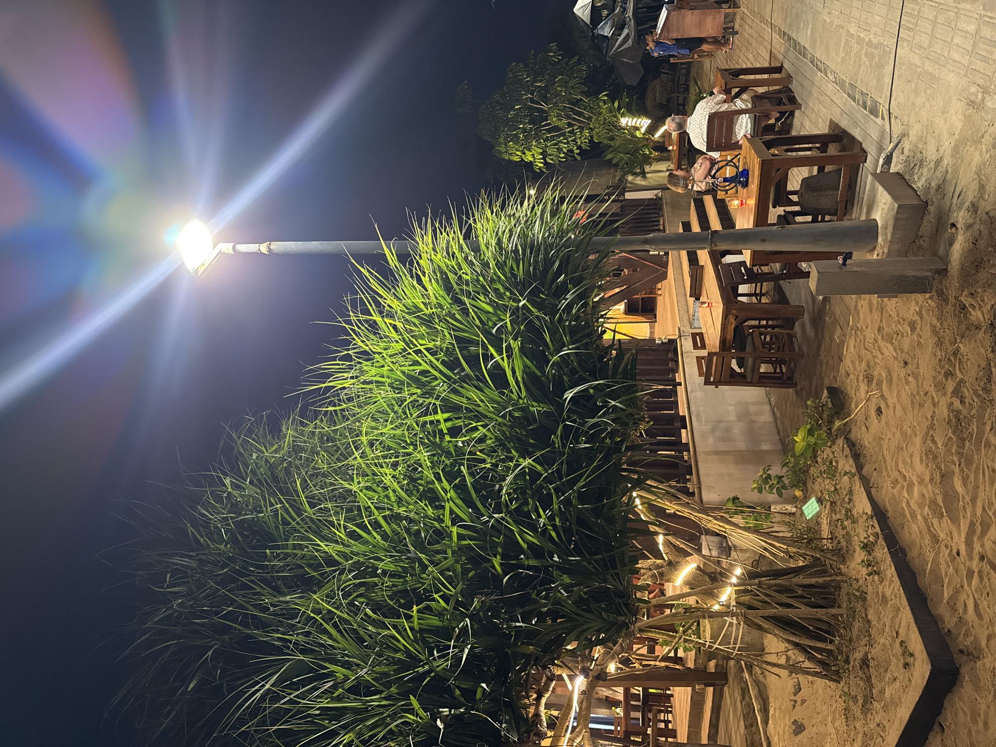 This image depicts an outdoor seating area at night, possibly a restaurant or bar, located on a sandy beach in Kuta, Indonesia. A large, lush green pandanus tree with exposed roots, decorated with string lights, dominates the left side of the frame. To the right, a pathway paved with rectangular stones runs parallel to the beach. Several wooden tables and chairs are arranged on a wooden deck overlooking the sand.
In the foreground, the sand is uneven and textured, with some scattered debris and small plants. A street lamp with a bright white light stands tall near the pandanus tree, casting beams of light and creating lens flare effects in the dark night sky.
On the right side of the image, a couple is seated at one of the tables, facing away from the camera. The woman has blonde hair and is wearing a patterned dress, while the man has short, light-colored hair and is wearing a patterned shirt. They appear to be engaged in conversation. Further back, another person in a blue shirt is standing near a tall wooden structure, possibly a counter or a menu display. Other tables and chairs are visible, some occupied by unseen patrons.
In the background, there are more trees and glimpses of buildings or structures, hinting at the urban environment of Kuta. The overall atmosphere is relaxed and inviting, suggesting a pleasant evening setting on the beach.
