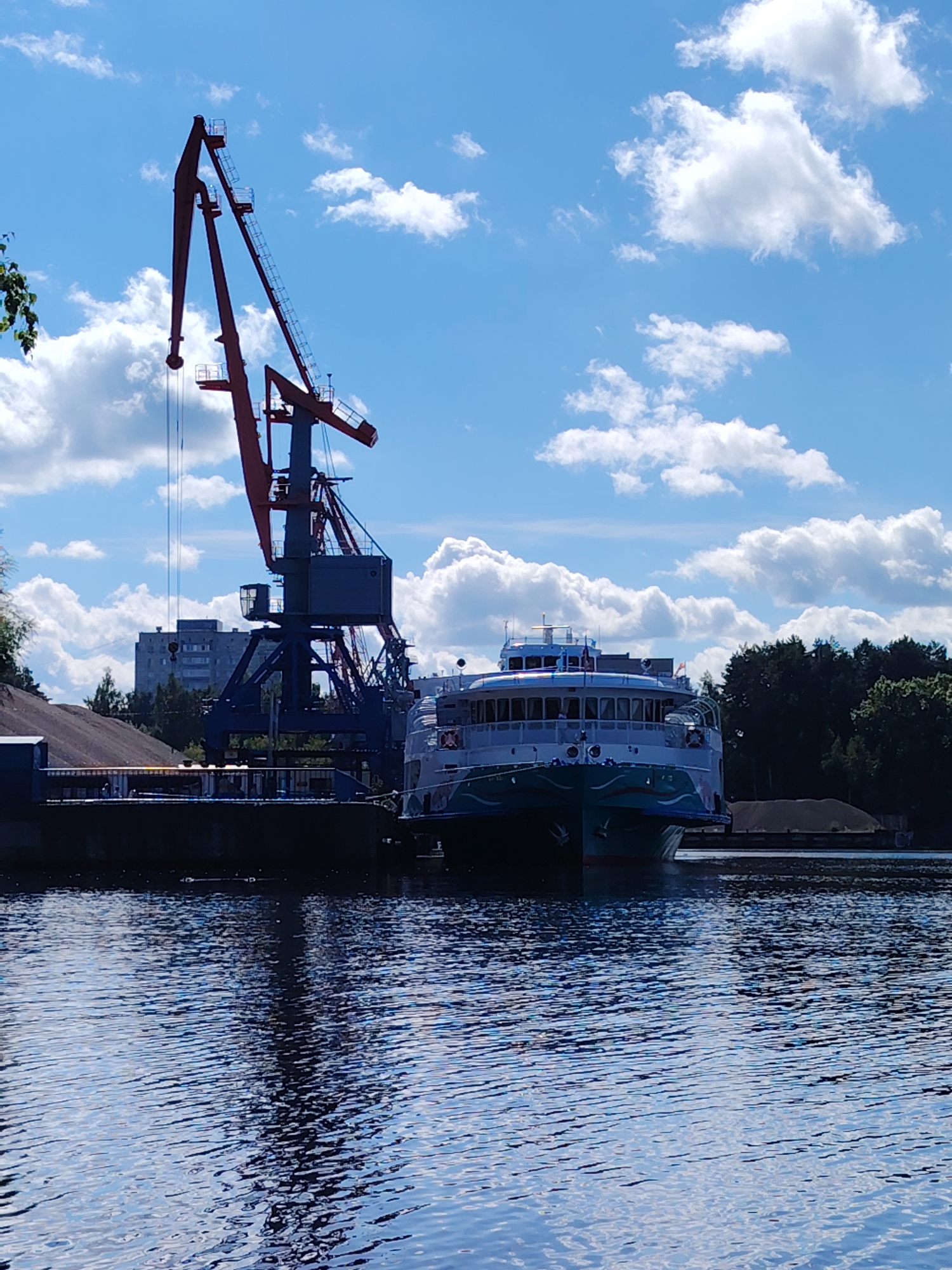 The image depicts a waterfront scene in the city of Chaikovsky, Russia. In the foreground, calm, blue water reflects the sky and surrounding objects. A large cargo ship, painted in white and teal with decorative blue patterns along its hull, is docked to the right. Behind the ship and to the left, a massive industrial crane with a red boom and blue structure stands tall against a bright blue sky dotted with fluffy white clouds. A multi-story building, likely an apartment complex, is visible in the background behind the crane. The shoreline features a dark embankment or dock area with a pile of sand or gravel. Trees form a dark green line along the far bank of the water. The overall impression is one of industrial activity and maritime operations on a sunny day.