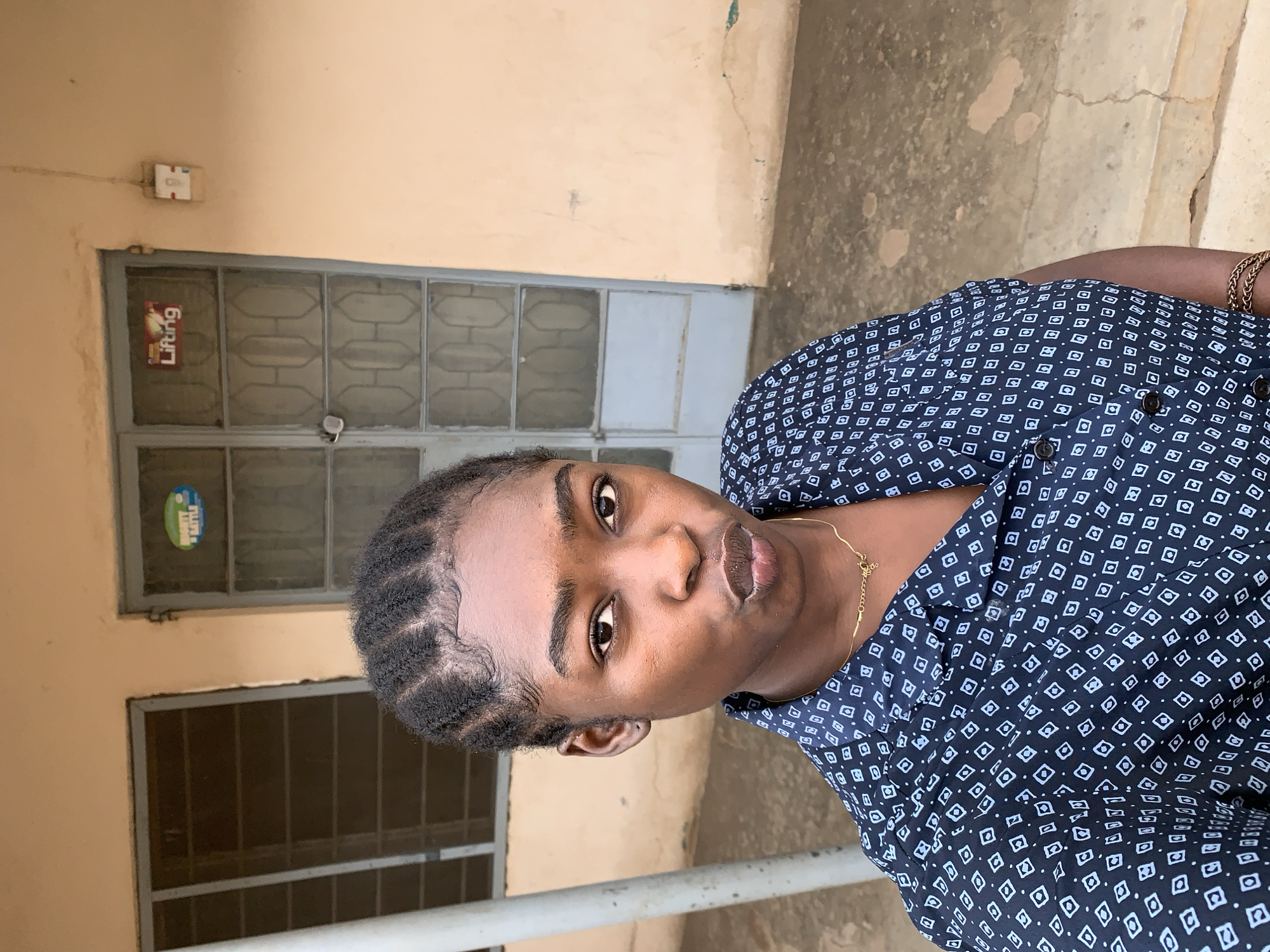 The image shows a young woman in Rafin Par, Nigeria. She has dark skin, braided hair, and is wearing a dark blue patterned shirt with buttons. She is also wearing a gold necklace. Behind her is a building with a window and a metal door with a "Lifting" sign on it. The ground appears to be concrete. The scene is illuminated by natural light.