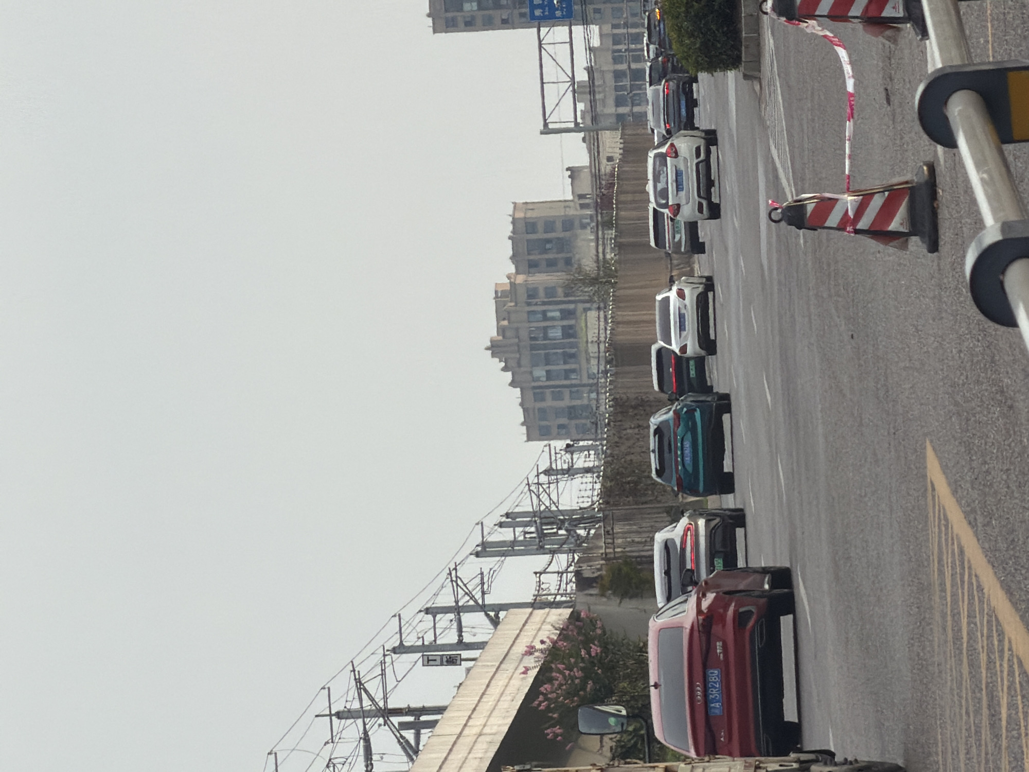 The image depicts a traffic scene on a road in Jiangjin, China, with multiple vehicles. In the foreground, a red Audi sedan with license plate "A 3R280" is visible. Further down the road, several other cars are in traffic, including a white sedan, a dark green SUV, and other vehicles in the distance. Above the road, an elevated structure with overhead power lines and electrical poles is present, suggesting it could be a railway or an elevated highway. In the background, a large, multi-story building complex with a distinctive architectural style is visible, likely part of the urban landscape of Jiangjin. The sky is overcast, and the overall lighting suggests it is daytime. Traffic cones and barrier tape are set up in the foreground, indicating some form of roadwork or traffic management.