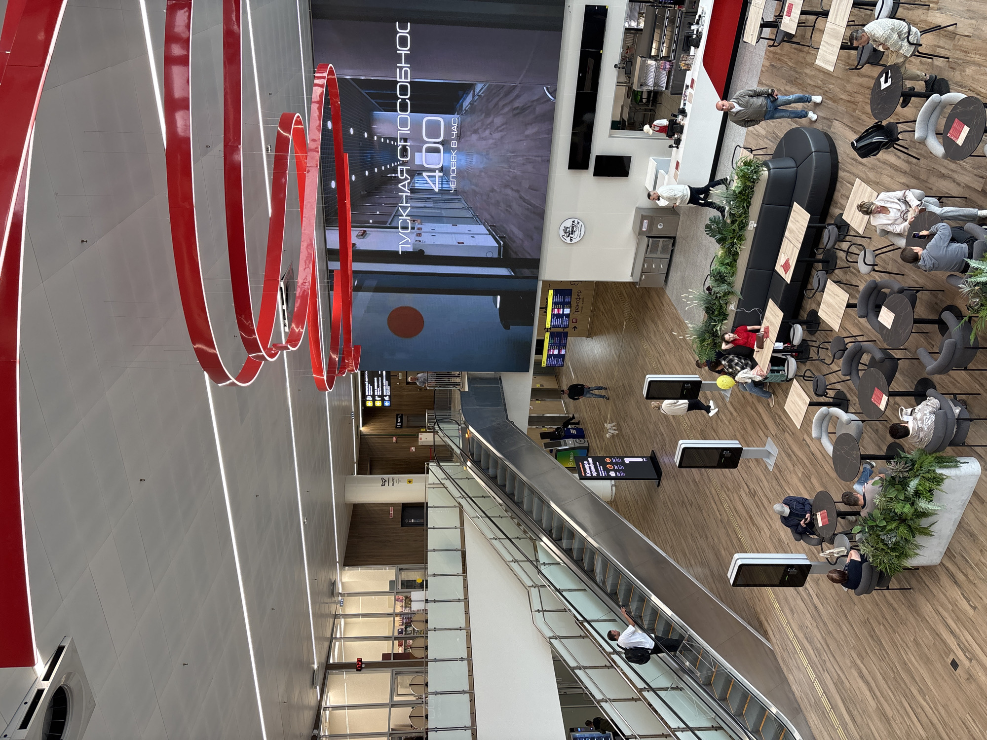 The image depicts an interior scene of a modern, spacious airport terminal, likely in Старое Мартьяново, Russia. The setting is characterized by polished wooden floors, a high ceiling with integrated lighting, and large screens displaying advertisements or information.
Several individuals are present, engaged in various airport-related activities. Some are seated at tables in a cafe or waiting area, others are standing or walking, and a few are using an escalator. The mood appears casual and relaxed, typical of a travel hub.
Notable objects include a large, prominent red spiral sculpture hanging from the ceiling, which serves as a striking visual element. There are also modern seating arrangements, including a long black curved sofa, individual chairs, and tables. Digital information screens are strategically placed throughout the terminal.
The large screen in the background displays Russian text, which translates to "TUKHNA. COMFORTABLE. 400 PEOPLE IN AN HOUR." This suggests it's an advertisement or an indicator of the airport's capacity. Other signs in the image also feature Russian text, indicating directions or services within the terminal.
The overall scene is well-lit, suggesting it is daytime or the terminal is brightly illuminated. No specific weather conditions are visible, as the image is entirely indoors. The activity is a general flow of passengers within an airport, with elements of relaxation and waiting.