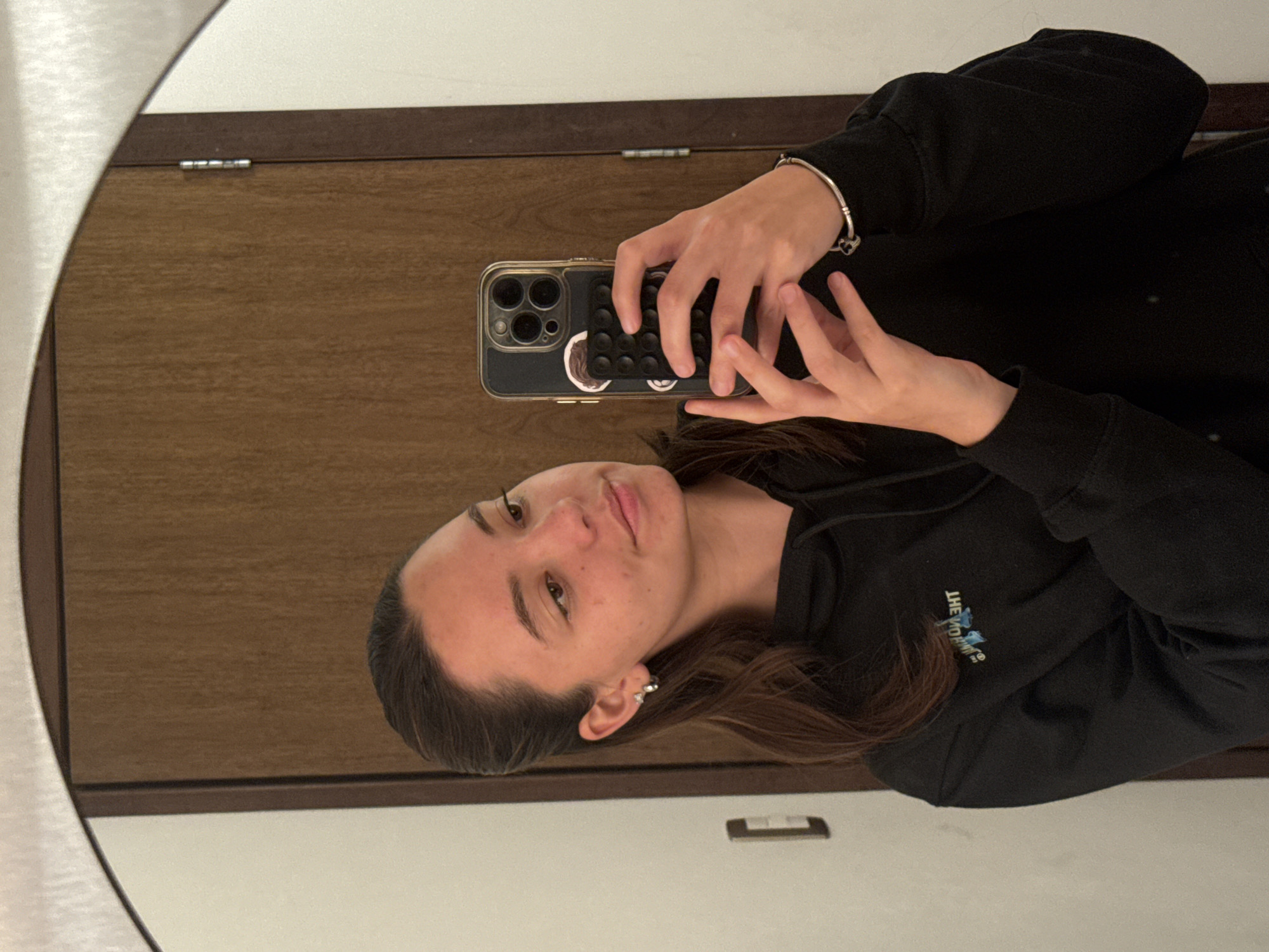 A young woman is taking a selfie in a mirror. She is wearing a black hooded sweatshirt and a silver bracelet on her left wrist. She holds a smartphone in both hands, the phone case features a black "pop-it" fidget toy attached to the back. The background consists of a wooden door or panel. The environment and lack of specific landmarks visible in the mirror make determining the location in San Bernardino Tlaxcalancingo, Mexico, impossible.