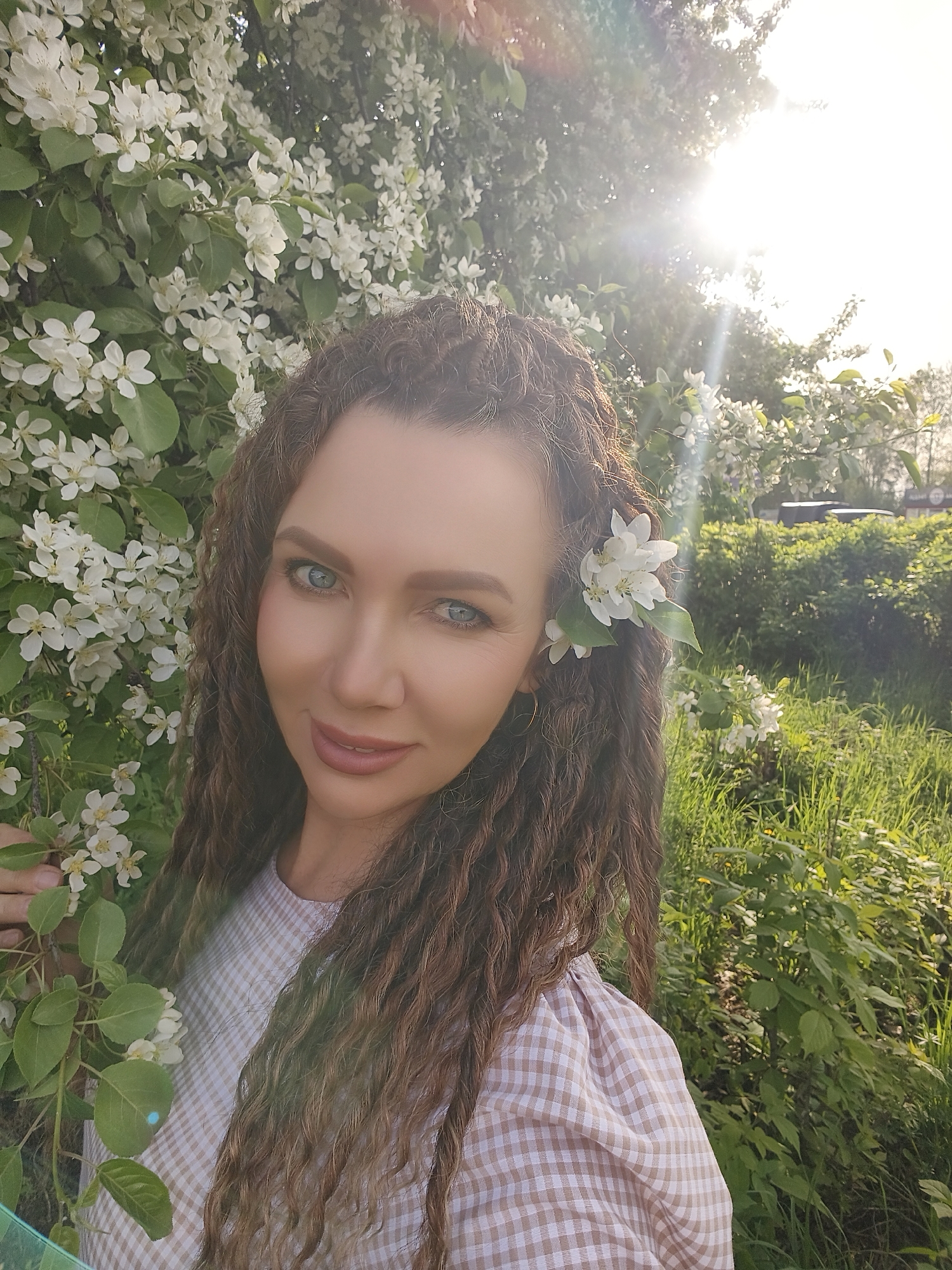 A woman with curly brown hair and blue eyes stands in a garden or park in Unknown, Unknown, surrounded by flowering trees and green foliage. A white flower is tucked behind her ear. She is wearing a tan and white checkered top. The sun is shining brightly, causing lens flare. The background includes a variety of plants, bushes, and grassy areas.
