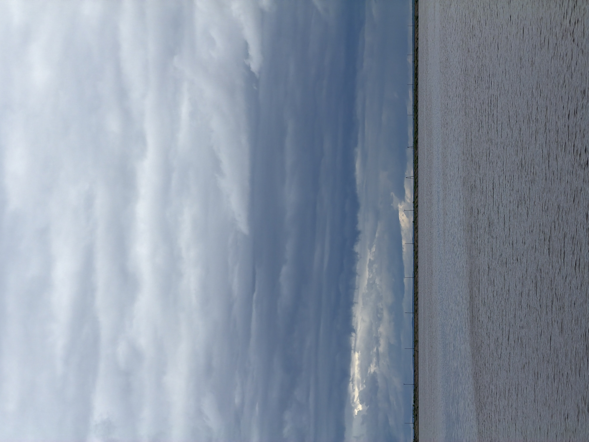 The image depicts a body of water under a cloudy sky. The water appears calm with subtle ripples. In the distance, there are utility poles along the shoreline. The location is Chaikovsky, Russia.
