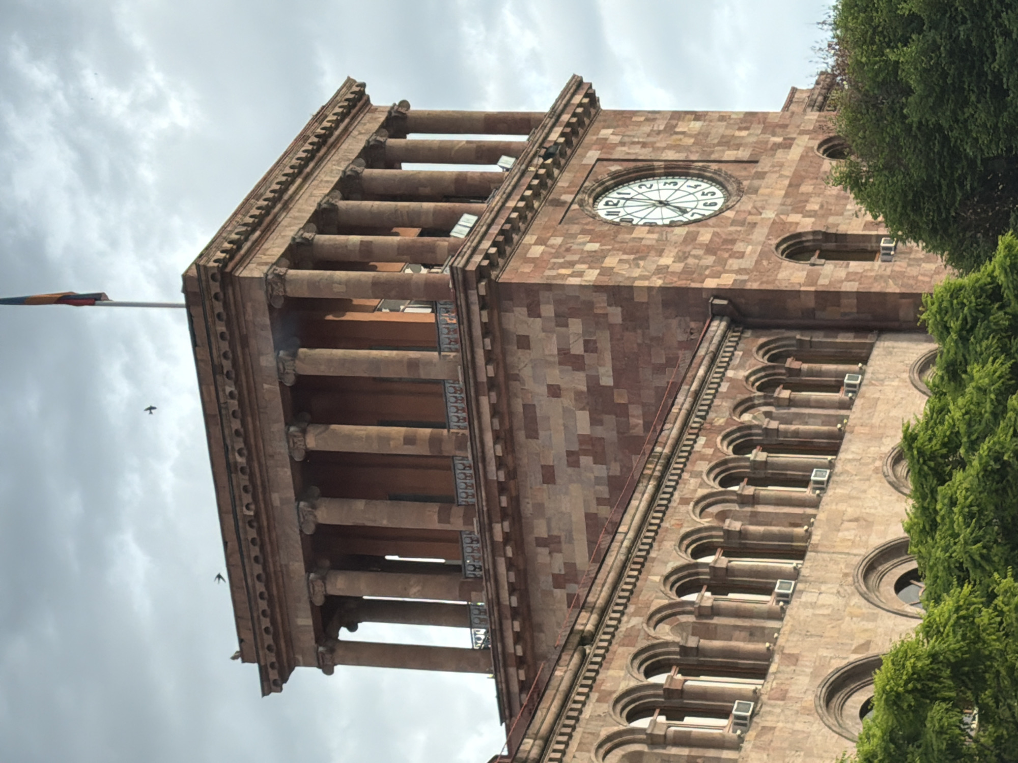 The image shows the architectural details of a building in Yerevan, Armenia, specifically the clock tower of the Yerevan City Hall. The building is constructed of brick and stone, featuring repeating patterns of arches, columns, and a clock face. A flag flies from a pole near the top of the building. Trees partially obscure the right side of the facade. The sky is overcast with gray clouds. Two birds are visible in the sky.