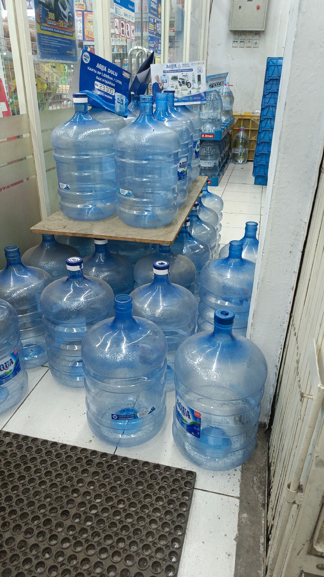 In the city of Margajaya, Indonesia, a retail store interior is depicted, showcasing a substantial display of bottled water. The scene is dominated by numerous large, clear plastic water jugs, predominantly filled with water and exhibiting the "AQUA" brand. These jugs are arranged in stacks on the floor and on a wooden shelf, with some partially obscured by others.
In the foreground, a dark, circular-patterned mat is visible on the tiled floor. In the background, merchandise is visible on shelves and racks, including what appears to be cleaning supplies or toiletries. Various promotional materials and signage are present on the walls and near the water jugs, some featuring prices and brand logos.
The lighting suggests an indoor environment, likely a convenience store or minimarket. No individuals are directly visible in the frame, though reflections in some of the bottles suggest the presence of other people in the store. The overall impression is one of organized inventory and product display.
