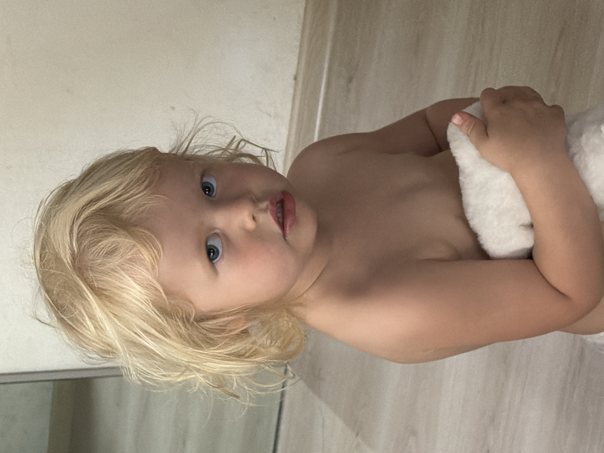 An eye-level portrait captures a young child, likely a toddler, in an indoor setting in Sochi, Russia. The child, with fair skin, bright blonde hair, and striking blue eyes, gazes directly at the camera with a curious and slightly open-mouthed expression, revealing some teeth. Their bare shoulders and upper torso are visible, suggesting they are shirtless or partially dressed. Both hands hold a soft, white, fluffy object, possibly a towel or a plush toy, against their chest.
The background is minimalist, consisting of light-colored walls and a light-toned wooden or tiled floor. The lighting appears soft and even, typical of an indoor environment, making the specific time of day indeterminate. There are no other discernible objects or specific activities taking place beyond the child posing for the photograph. No visible text is present in the image. The overall impression is one of candidness and innocence.