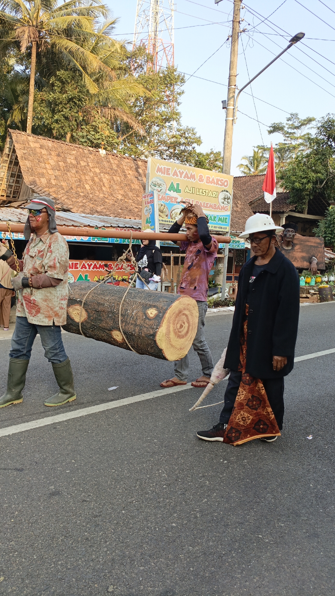 In Krajan Lor, Indonesia, a group of people are participating in a procession, carrying a large, artificial log across a paved road. The log is made to resemble wood, with painted rings and bark texture, and is suspended from a wooden pole supported by two men. The man on the left is wearing green rubber boots, jeans, and a stained, long-sleeved shirt. He also has sunglasses and face paint. The man on the right, carrying the other end of the pole, is wearing a colorful, ripped t-shirt and flip-flops, with a patterned headwrap. To the right of them, an older man in a black jacket and white hat walks alongside, with a patterned fabric draped over his arm. In the background, a building with a tiled roof and a prominent sign advertising "MIE AYAM & BAKSO AL AJI" is visible, along with trees and a cell tower. A red and white Indonesian flag is also present. Other individuals, including women in headscarves, are seen in the background, suggesting a community event or parade. The scene is captured in daylight, with shadows indicating the sun is relatively low in the sky.