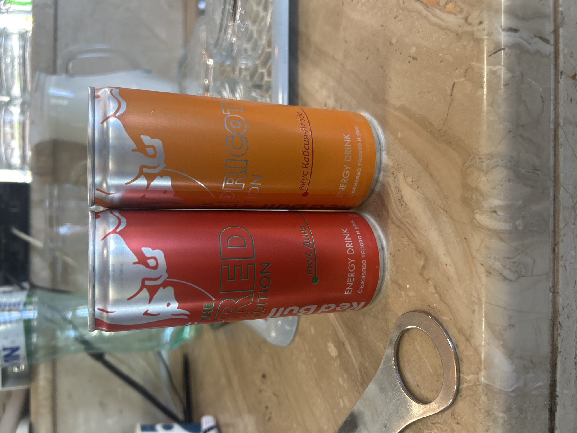 On a marble countertop, two cans of Red Bull energy drink are stacked. The top can is orange and labeled "Apricot Edition" in English, with additional text in Cyrillic. The bottom can is red and labeled "Red Edition" in English, also with Cyrillic text. A silver bottle opener lies next to the cans. Background elements include glassware and a partially visible cabinet. The cans are new and seem to be a common type in Kazanlak, Bulgaria.