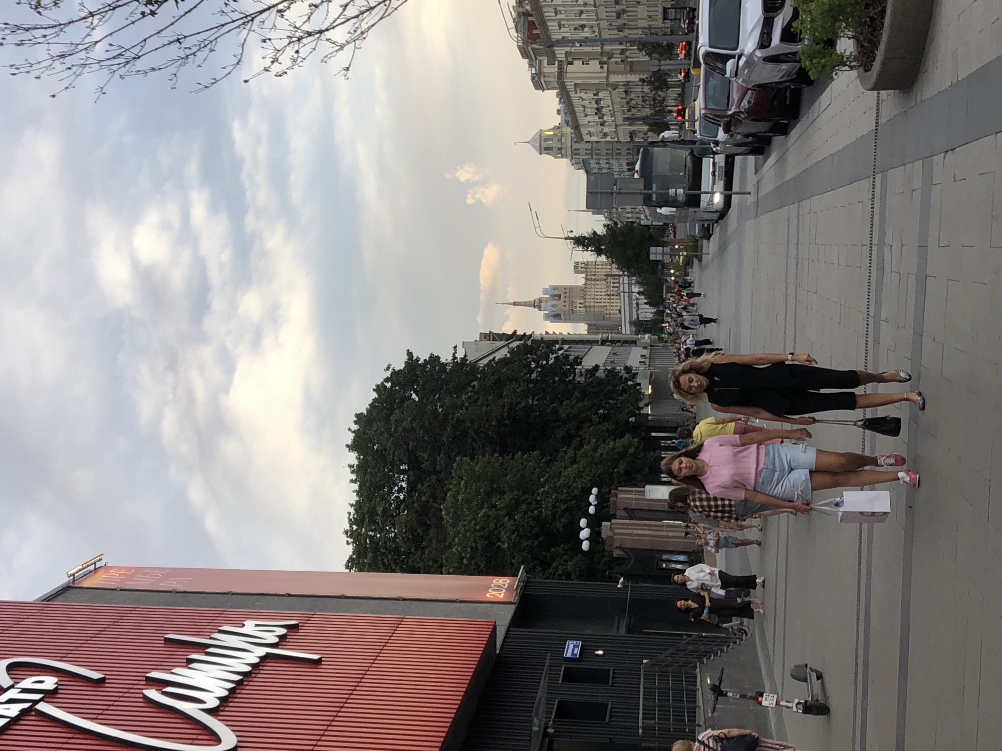 The image shows a street scene in Moscow, Russia, likely in the evening. A large red building with white lettering on its side is on the left, possibly a theater or cultural center. The lettering is in Russian script and appears to spell "Театр Сатиры" which translates to "Satire Theatre". The building has vertical red ribbed panels. Further down the street, people are walking on a paved sidewalk. A bus and several cars are parked or driving on the street. In the background, there are buildings with distinct architectural styles, including a tall, ornate skyscraper with spires. The sky is cloudy, with hints of sunset colors. Two women are walking towards the camera in the foreground; one is wearing a pink top and denim shorts and carrying a shopping bag, while the other is wearing a black outfit and carrying a handbag. There are several other pedestrians visible on the sidewalk. An electric scooter is parked near the building on the left.