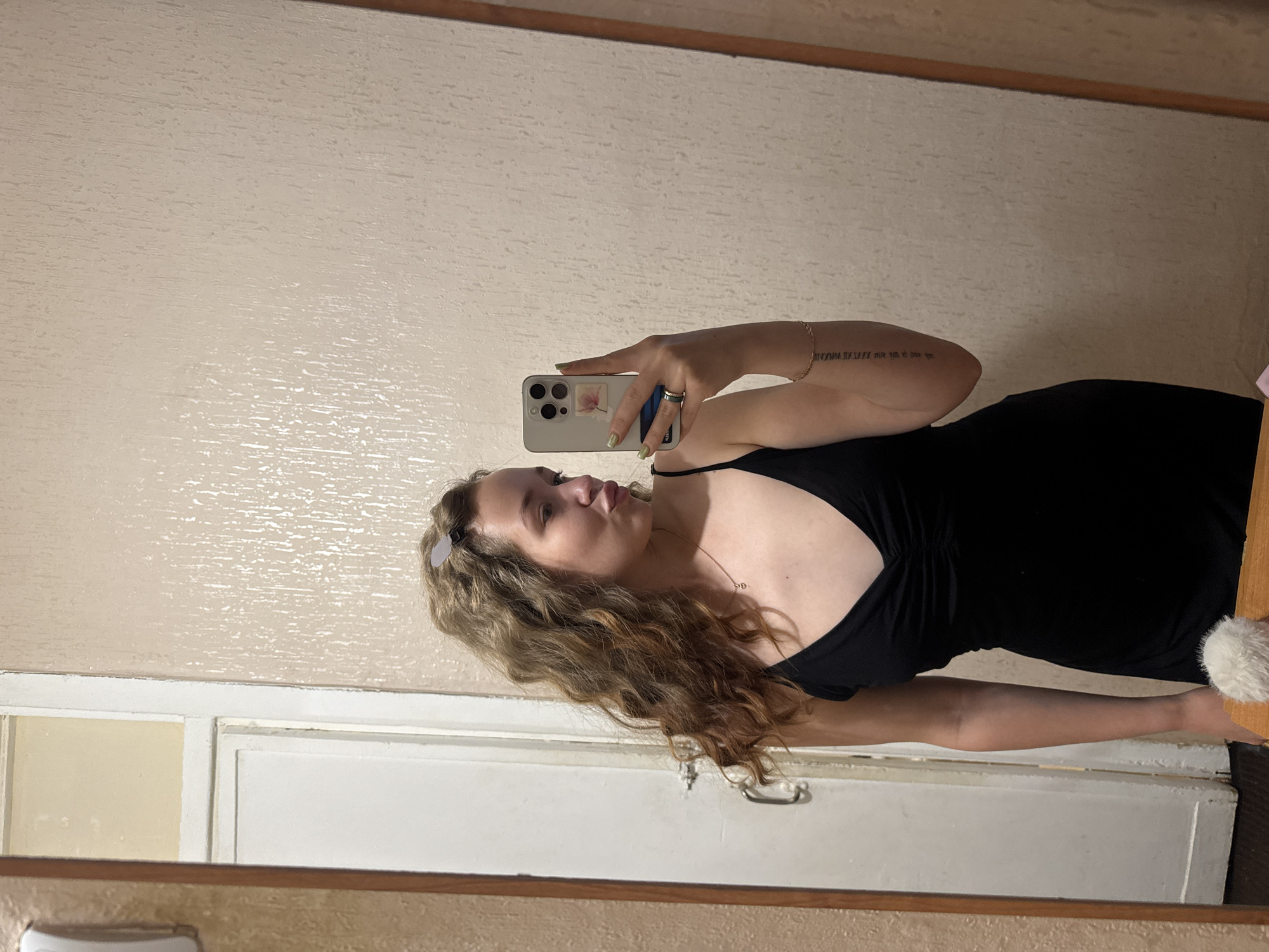 In a dimly lit room, a young woman with curly blonde hair takes a mirror selfie. She is wearing a black spaghetti strap dress and has a playful pout. Her left hand, holding the phone, shows off a light green nail polish and a ring. A delicate gold chain with a small heart pendant is visible around her neck. A subtle tattoo runs vertically along her left forearm. Behind her, a white door with a silver handle is partially visible, along with textured beige wallpaper. The setting appears to be a residential interior, possibly an apartment in Chaikovsky, Russia, given the typical architecture and decor associated with the region.
