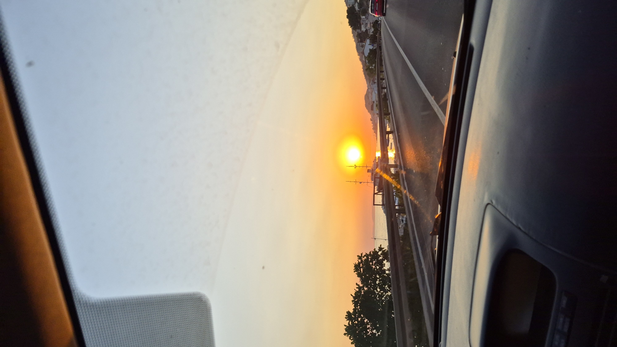 The image is taken from inside a car looking out the windshield at a sunset. The sun is low in the sky, casting a warm orange glow over the scene. There is a body of water, likely the sea, with a ship in the distance. The road ahead is paved and has white lane markings. To the left of the road, there is a metal guardrail and some trees. To the right of the road, there are buildings on a hillside. The interior of the car, including the dashboard, is visible in the foreground. The sky is a gradient of orange and blue.