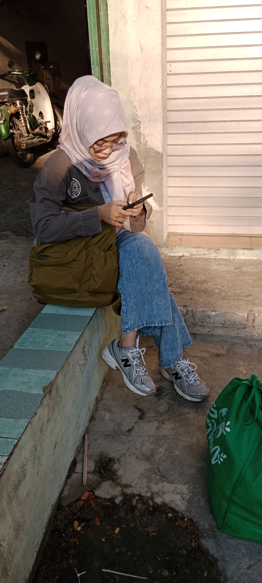 A young woman wearing a light purple hijab and glasses is sitting on a concrete ledge outdoors, holding and looking at her phone. She is dressed in a grey jacket over a striped shirt and loose-fitting blue jeans, with silver athletic shoes on her feet. A large olive green messenger bag is slung over her left shoulder and rests on the ledge beside her. To the right of the frame, a bright green drawstring bag with a white floral pattern is visible. The background shows a section of a building with white horizontal shutters and a partially visible motorcycle parked in an open doorway to the left. The scene appears to be in an urban setting, possibly Bojonegoro, Indonesia, given the architectural style and common presence of motorcycles. The lighting suggests it is late afternoon or early morning, with warm sunlight casting shadows.