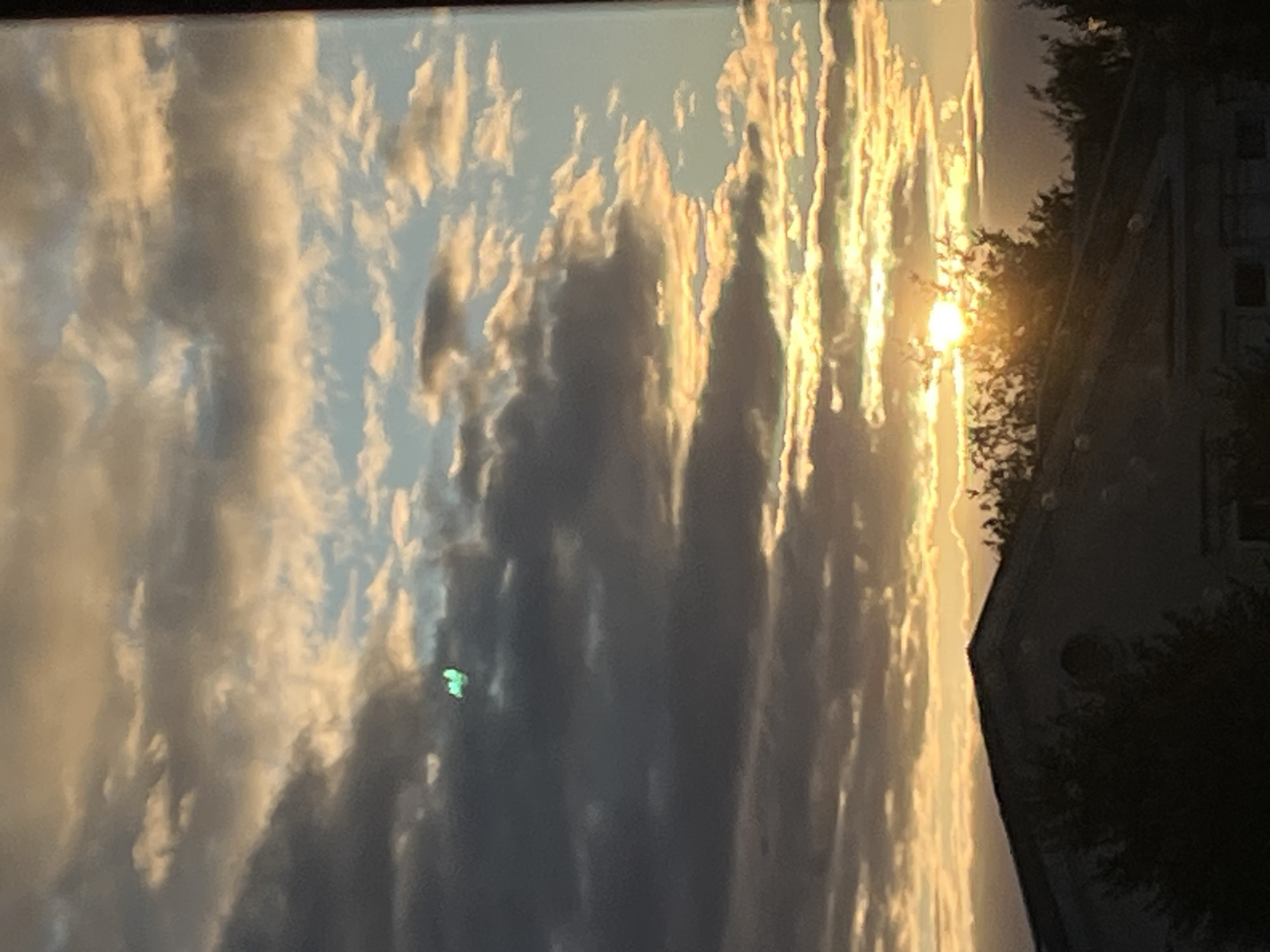 The image captures a sunset over a cloudy sky. The sun's rays break through the clouds, creating streaks of golden light. On the right side of the image, the dark silhouette of a building and trees can be seen, suggesting an outdoor setting. The overall mood is serene and peaceful, characteristic of a sunset. The time of day is clearly evening, given the setting sun. The clouds suggest it might be partly cloudy weather. There is a small, bright green object visible in the clouds on the left side of the image. No text is visible. The provided location context is Kohtla-Järve linn, Estonia.