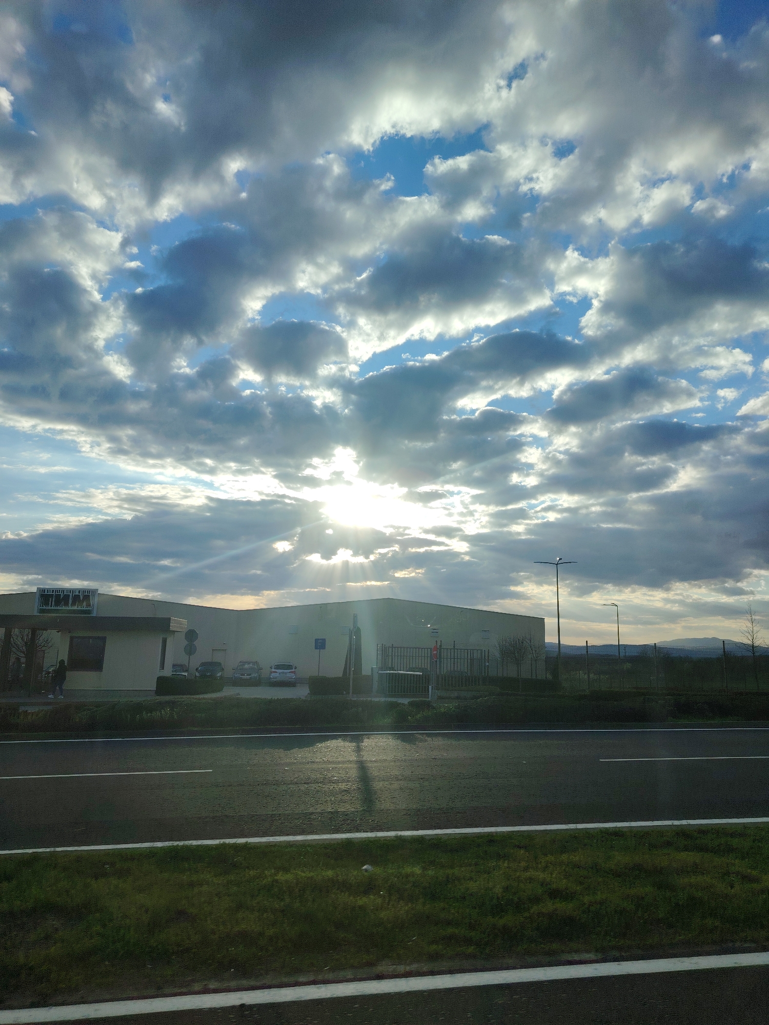 The image shows a view from a road in Lyaskovets, Bulgaria, towards an industrial area under a cloudy sky with the sun shining through the clouds. In the middle ground, there is a road with green grass and a white stripe on the bottom. Beyond the road, there is a complex of industrial buildings, a parking lot with cars, a fence, some vegetation, and a mountain range in the distance. A person walks on the left of the image near the building. Light poles are visible.