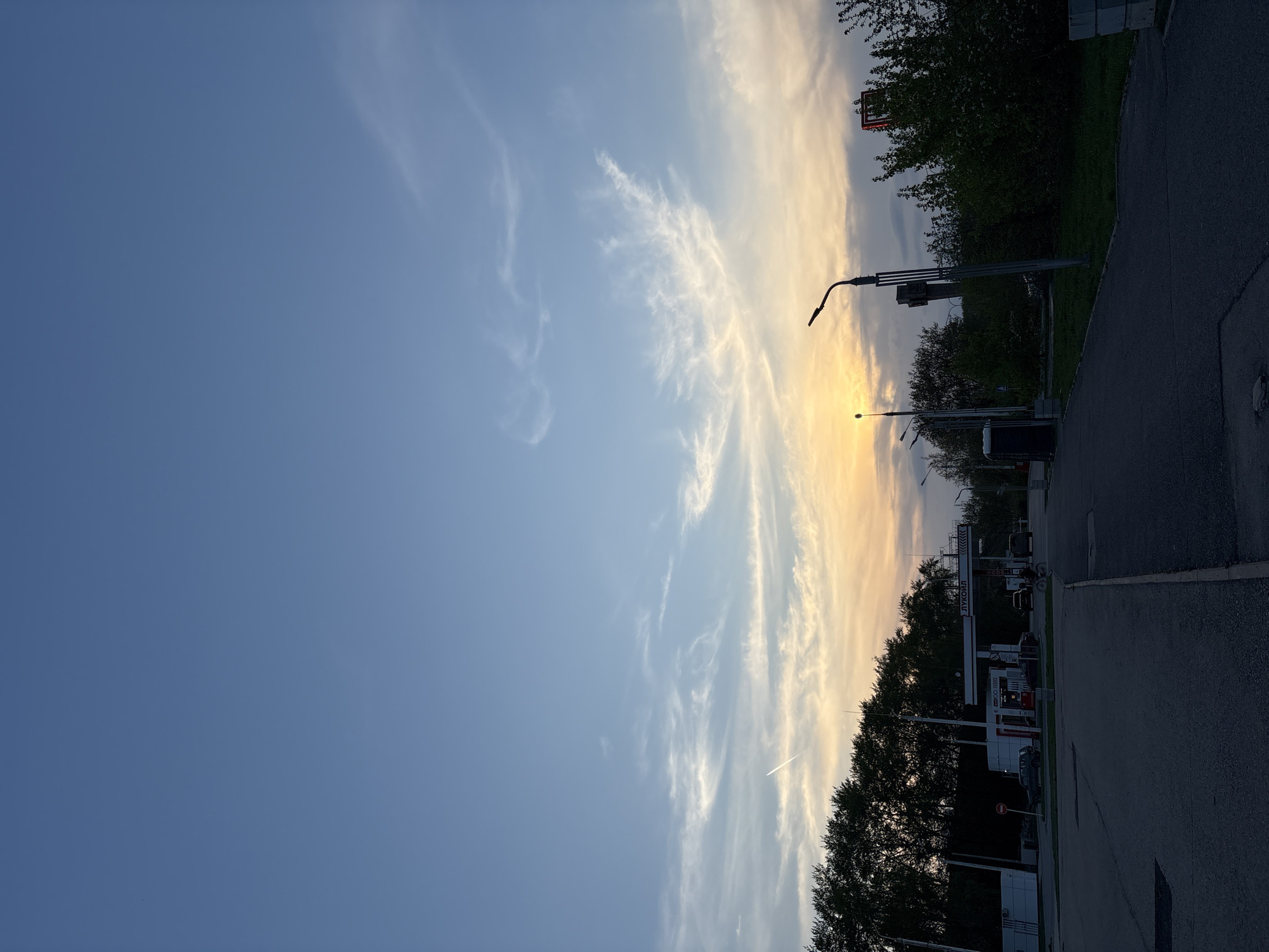 The image shows a street scene at dusk in Perm, Russia. The sky is a gradient of blue to yellow-orange, with wispy clouds. A Lukoil gas station is visible on the right side of the frame, with several cars parked near the pumps. A paved road runs along the right edge of the image. Streetlights line the road. Trees are planted between the road and the gas station. A contrail stretches across the middle of the sky.